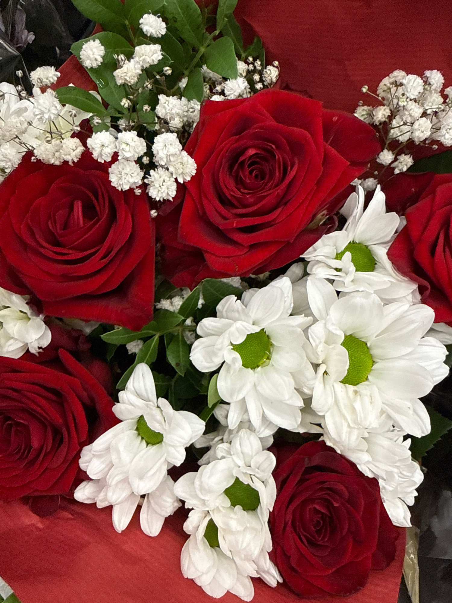 A close-up, eye-level shot shows a bouquet of red roses and white daisies with green centers. Small white baby's breath flowers are interspersed throughout the arrangement. The bouquet is presented against a red background, likely wrapping paper or fabric. The image is shot in Göttingen, Germany.