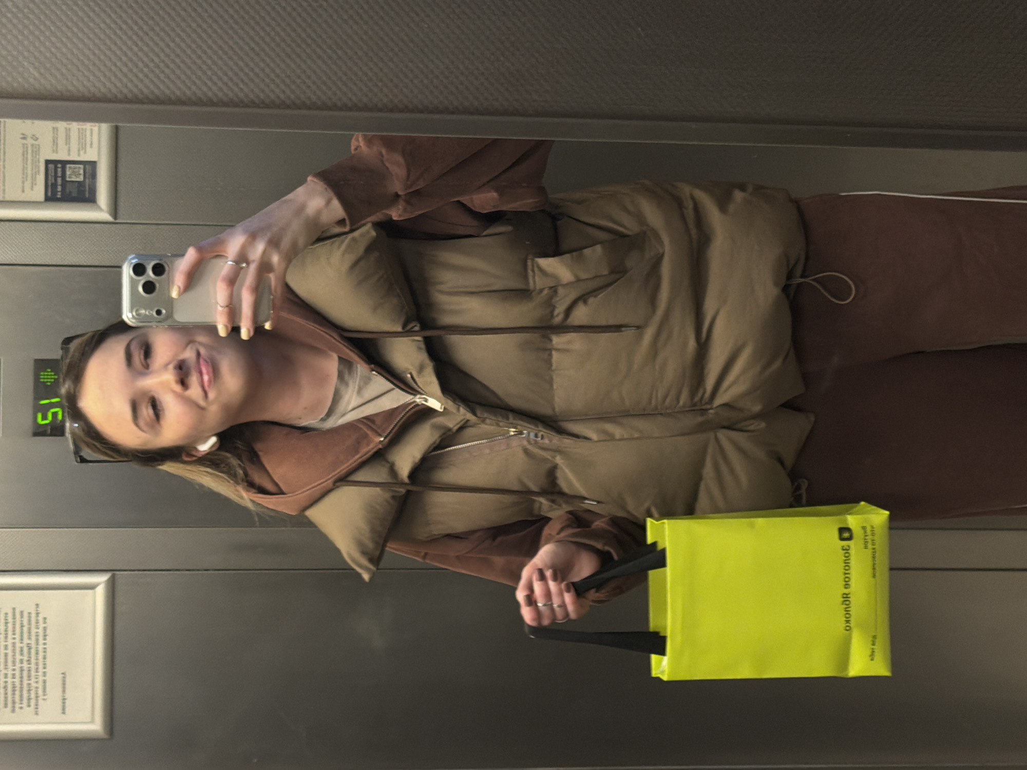 A young woman takes a selfie in an elevator. She wears a brown tracksuit with a beige puffer vest. She holds a bright yellow shopping bag with Russian text on it. The elevator displays the floor number 15. Text in Russian is visible on the elevator wall. The location is assumed to be within Kuleshovka, Russia.