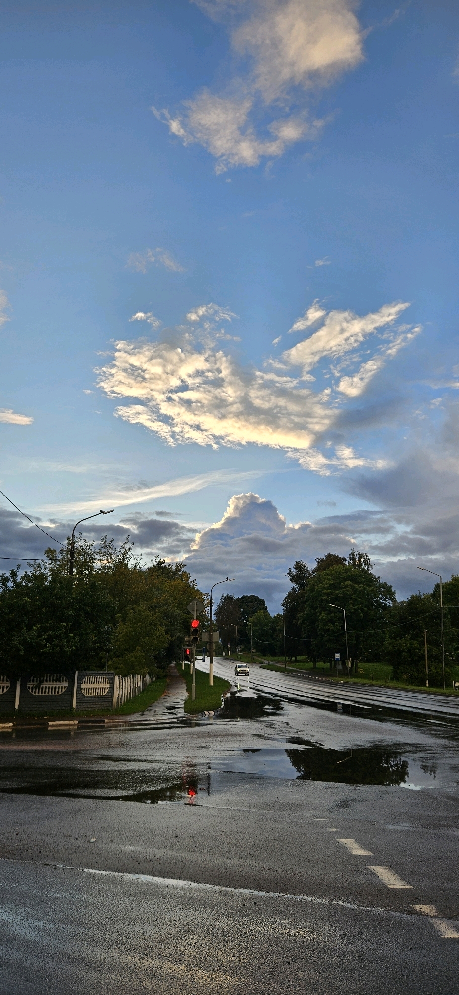 The image depicts a wet asphalt road in Vitsebsk, Belarus, likely after a rain shower. The sky is partly cloudy with white and grey clouds illuminated by sunlight, suggesting it might be late afternoon or early evening. A traffic light shows a red signal, indicating that vehicles should stop. There is a single white car on the road, stopped at the red light. On the left side of the road, there is a sidewalk with a low decorative fence and green grass. Tall trees with dense foliage line both sides of the road. Puddles of water on the road reflect the sky and the traffic light. In the distance, a large cumulonimbus cloud formation is visible, adding dramatic contrast to the sky. The overall scene conveys a sense of calm and quiet, with the reflective surfaces of the wet road creating an atmospheric effect.