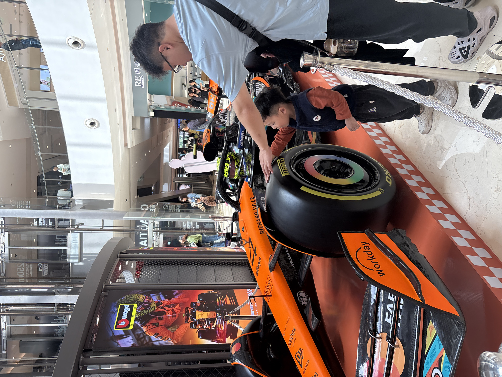 The image shows a man and a child looking at an orange McLaren Formula 1 car display in a mall. The man, wearing glasses and a light blue shirt, is touching the tire of the race car. The child, wearing an orange shirt and navy vest, is also looking at the tire. The car is displayed on a platform with a checkered pattern and is surrounded by a rope barrier. In the background, people can be seen walking in the mall. The display has a large screen showing a race car graphic. Logos of "burago", "CISCO", "CASTORE", "webex", "Hilton", "RICHARD MILLE", and "McLaren" are visible on the car and display. The mall has multiple levels with shops and glass panels. The environment suggests a modern shopping center. Based on the OCR result, the location is likely within Yuzhong District, China, as suggested by the term "RE调香 CLASSIFIED" and the mention of "Aren LA 1 TEAM".