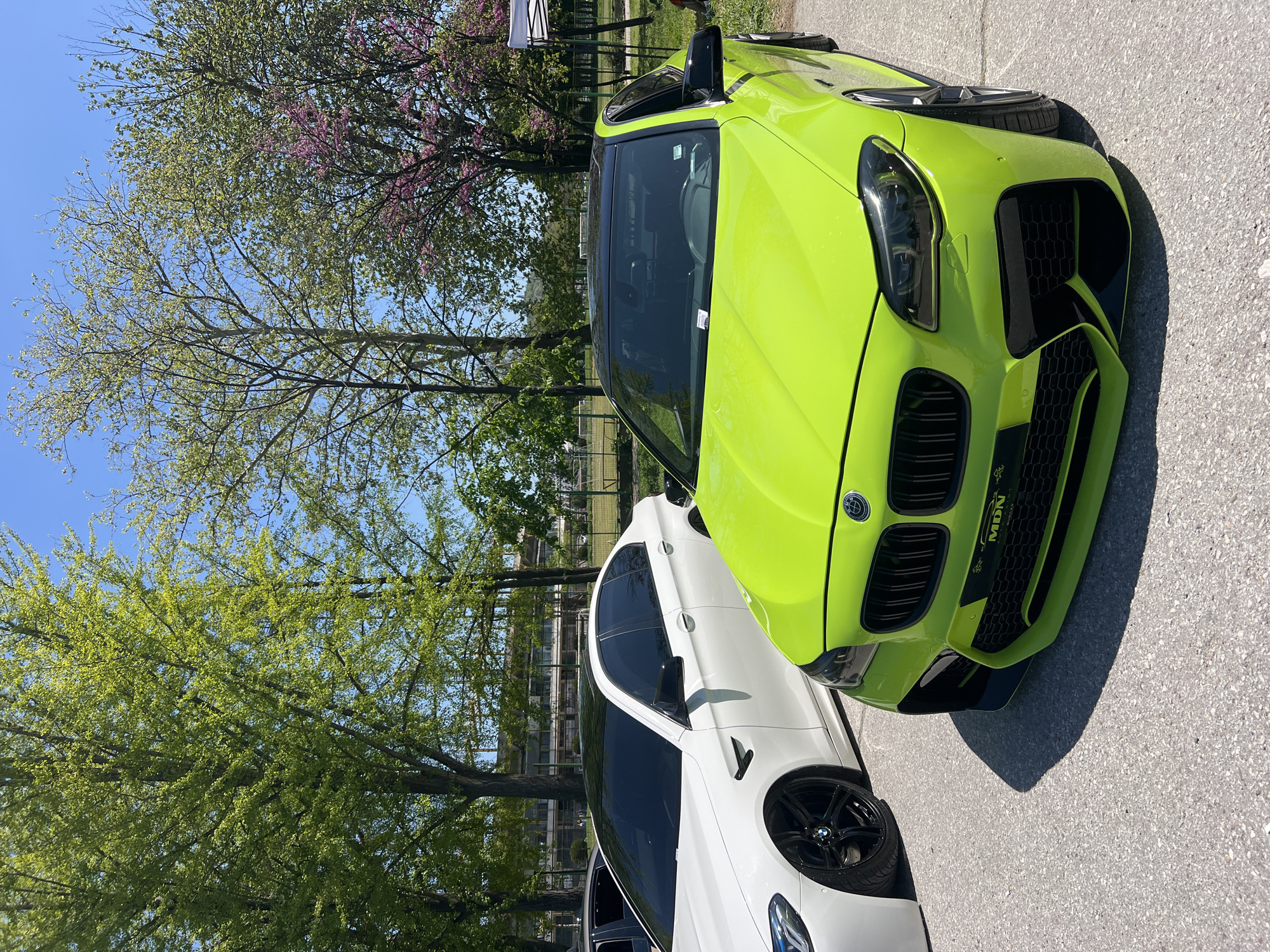 The image depicts two cars parked on an asphalt surface. The car in the foreground is bright lime green with a black grill. The car is angled towards the viewer. A white car is visible next to the lime green car. The location is Plovdiv, Bulgaria. Trees with fresh green leaves are visible in the background, with some trees blooming with pink flowers. The sky is clear blue. A chain-link fence and partial building are in the background, hinting at a park or recreational area.