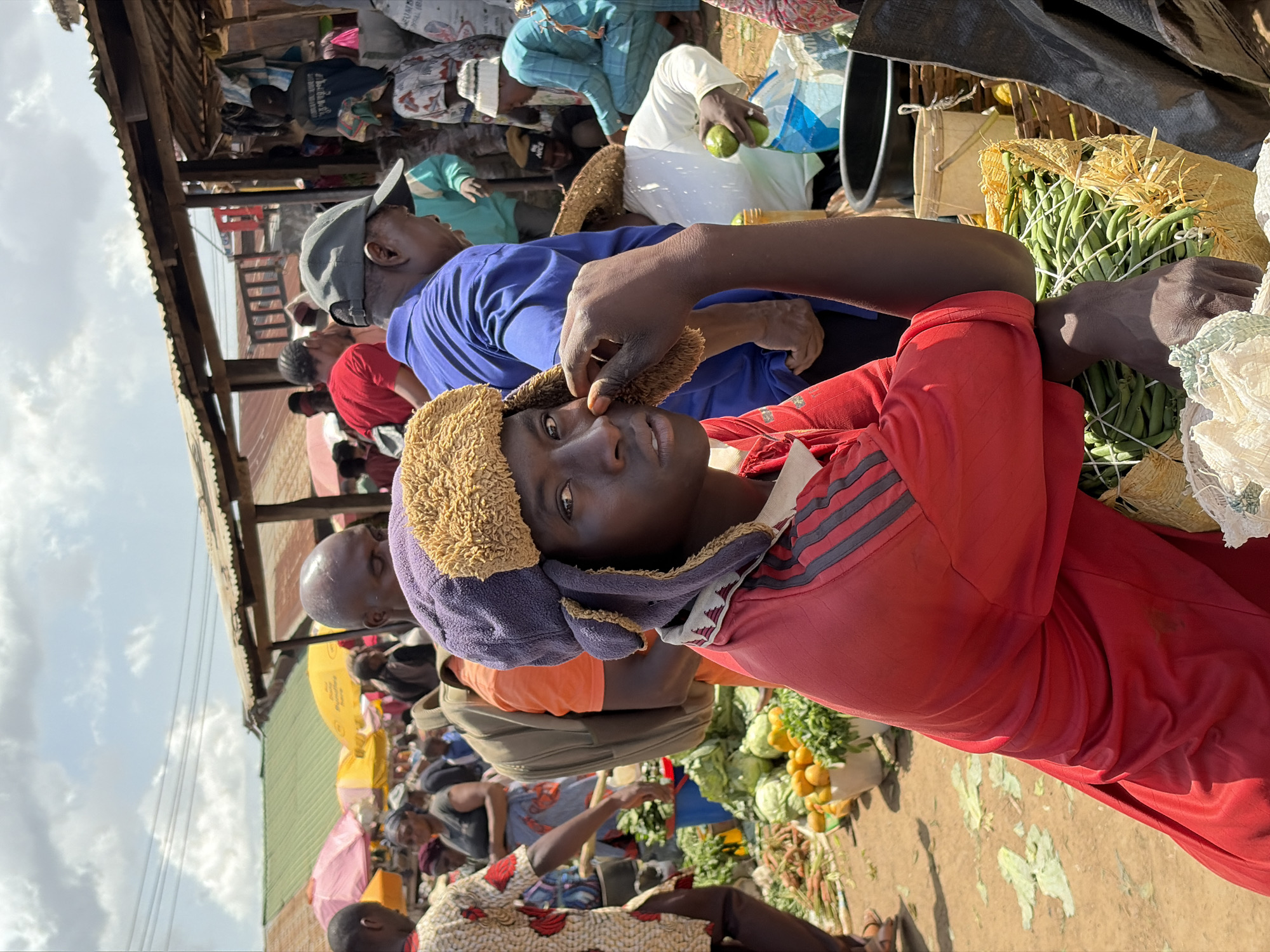 This media file captures a vibrant, crowded outdoor market scene in Gura Top, Nigeria, likely during daylight hours given the bright illumination and visible sky. The image is taken from a low perspective, looking upwards into the market activity.
The most prominent subject is a young individual in the foreground, wearing a red long-sleeved shirt with dark stripes on the shoulders and a distinctive, light brown furry hat with purple ear flaps. This person looks directly at the camera with a calm, neutral expression. An arm, likely belonging to another person, is visible near their face, possibly interacting with their hat.
Behind the foreground figure, the market is bustling with activity and numerous people. Many individuals are present, some engaged in transactions or browsing, while others move through the narrow passages between stalls. Notable figures include a man in a blue shirt and grey baseball cap looking upwards, and another person in a white outfit holding a green fruit, possibly an avocado.
The market features numerous stalls with makeshift structures, characterized by corrugated iron roofs providing shade. A diverse array of fresh produce is displayed, including large bundles of green beans, often tied with straw, as well as green cabbage and piles of yellow, round fruits. The ground appears to be unpaved dirt, with scattered vegetable debris. In the background, more people and stalls are visible, some shaded by colorful umbrellas, suggesting a typical market environment. The sky above is bright with visible clouds, indicating an overcast but well-lit day. No significant text is clearly legible within the frame.