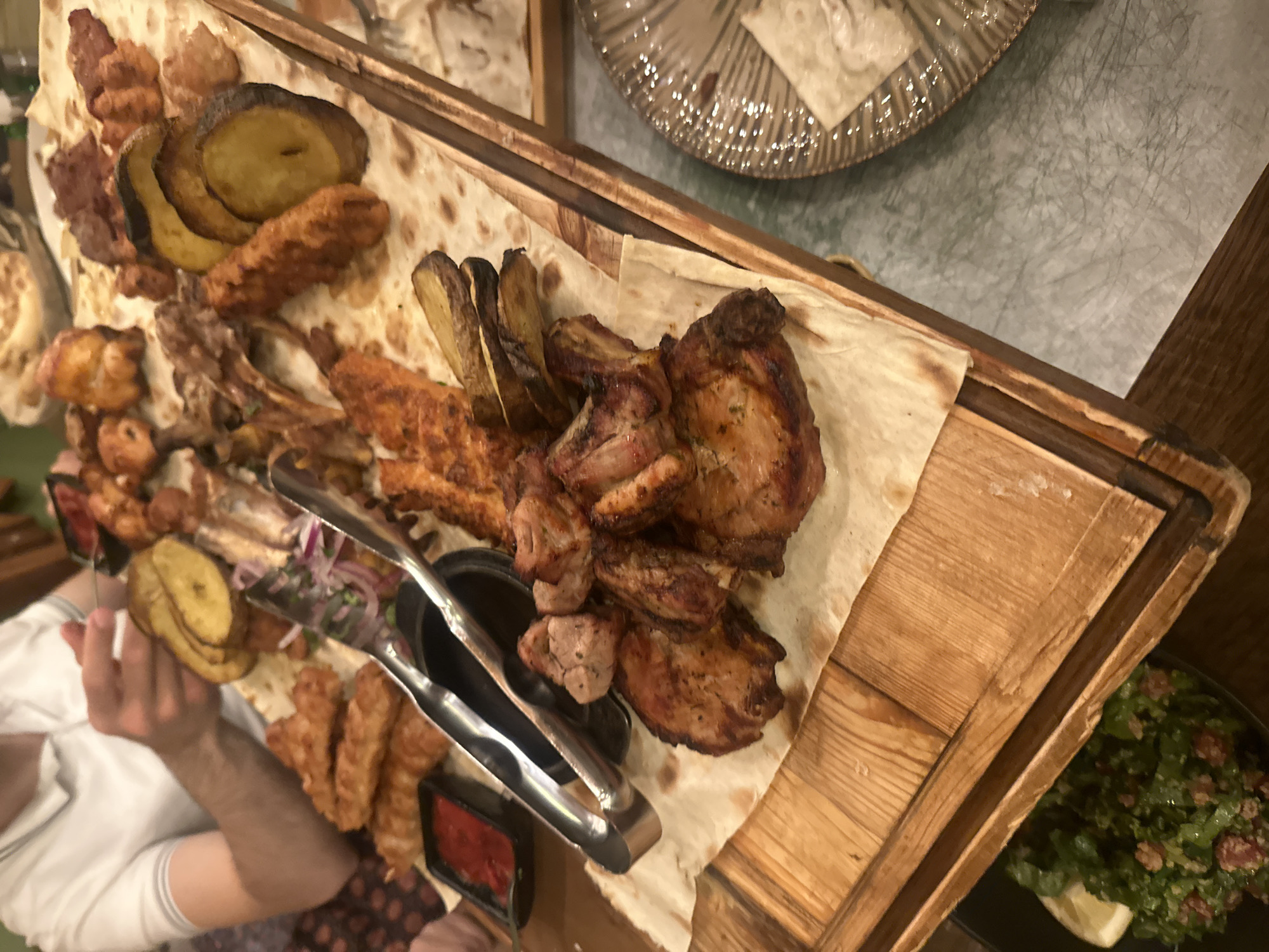 A generous platter of grilled meats and vegetables, characteristic of Armenian cuisine, is the central focus of this indoor dining scene. The large wooden rectangular board is laden with a variety of cooked items, including dark-browned chicken pieces, numerous skewers of what appear to be khorovats (Armenian BBQ) and lula kebab, and slices of grilled potatoes or eggplant. These items are presented on top of several pieces of lavash bread, which lines the platter and absorbs the juices.
To the left, a person's arm, wearing a white short-sleeved shirt with faint grey stripes, is visible, reaching towards the food, suggesting they are in the process of serving or eating. Metal tongs rest on the platter, near a small black bowl containing a vibrant red sauce, likely adjika or a similar tomato-based condiment. In the lower right corner, a portion of a black plate holds a fresh green salad, possibly tabbouleh, garnished with a lemon wedge.
The setting appears to be a restaurant or a casual dining establishment in Yerevan, Armenia. The lighting is warm and artificial, suggesting an evening meal. In the upper background, part of a decorative, textured plate or bowl is visible on a light-colored table surface, which has a subtly scratched texture. The overall presentation emphasizes abundance and communal dining, typical for such traditional meals. No visible text is present in the image.