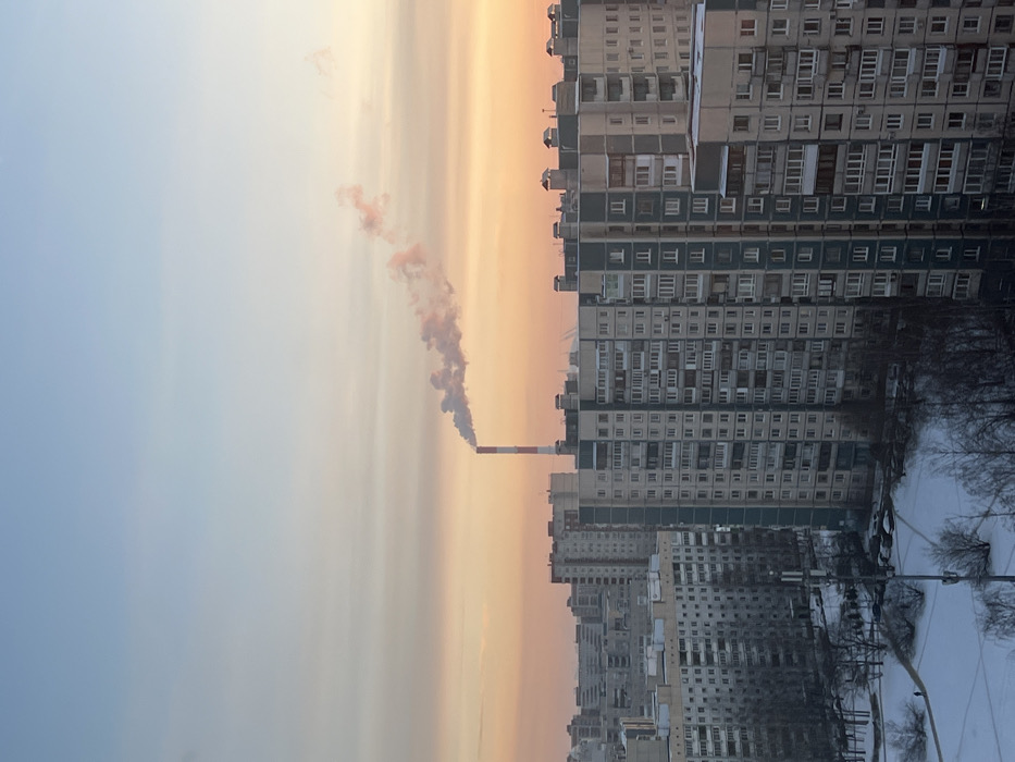 A view of Soviet-era apartment buildings is seen against a warm, orange sunset. A large smokestack emits a plume of smoke that blends into the colorful sky. Snow covers the ground and bare trees in the foreground. The apartments are uniform and repetitive, with rows of windows reflecting the setting sun. The image captures a blend of industrial activity, urban landscape, and natural beauty.