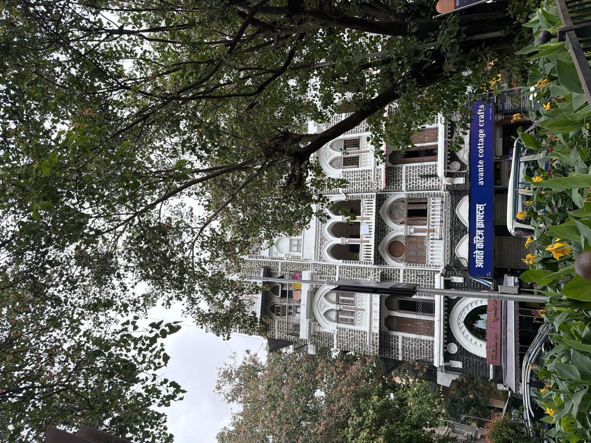 The image captures a street view in Mumbai, India, focusing on a multi-story building with distinct architecture, likely from the early 20th century, characterized by its stone facade, white arched window frames, and balconies. The building is partially obscured by lush green trees in the foreground and on the right side. Below the building, a row of shops is visible, including one with a blue sign that reads "Avante Cottage Crafts" in English and some text in Devanagari script. Cars are parked along the street, and yellow flowers are blooming in the foreground. The sky is overcast, indicating a cloudy day. The architectural style of the building, the presence of shops like "Avante Cottage Crafts," and the general street scene are characteristic of areas like South Mumbai, which retains many historical buildings from the colonial era.