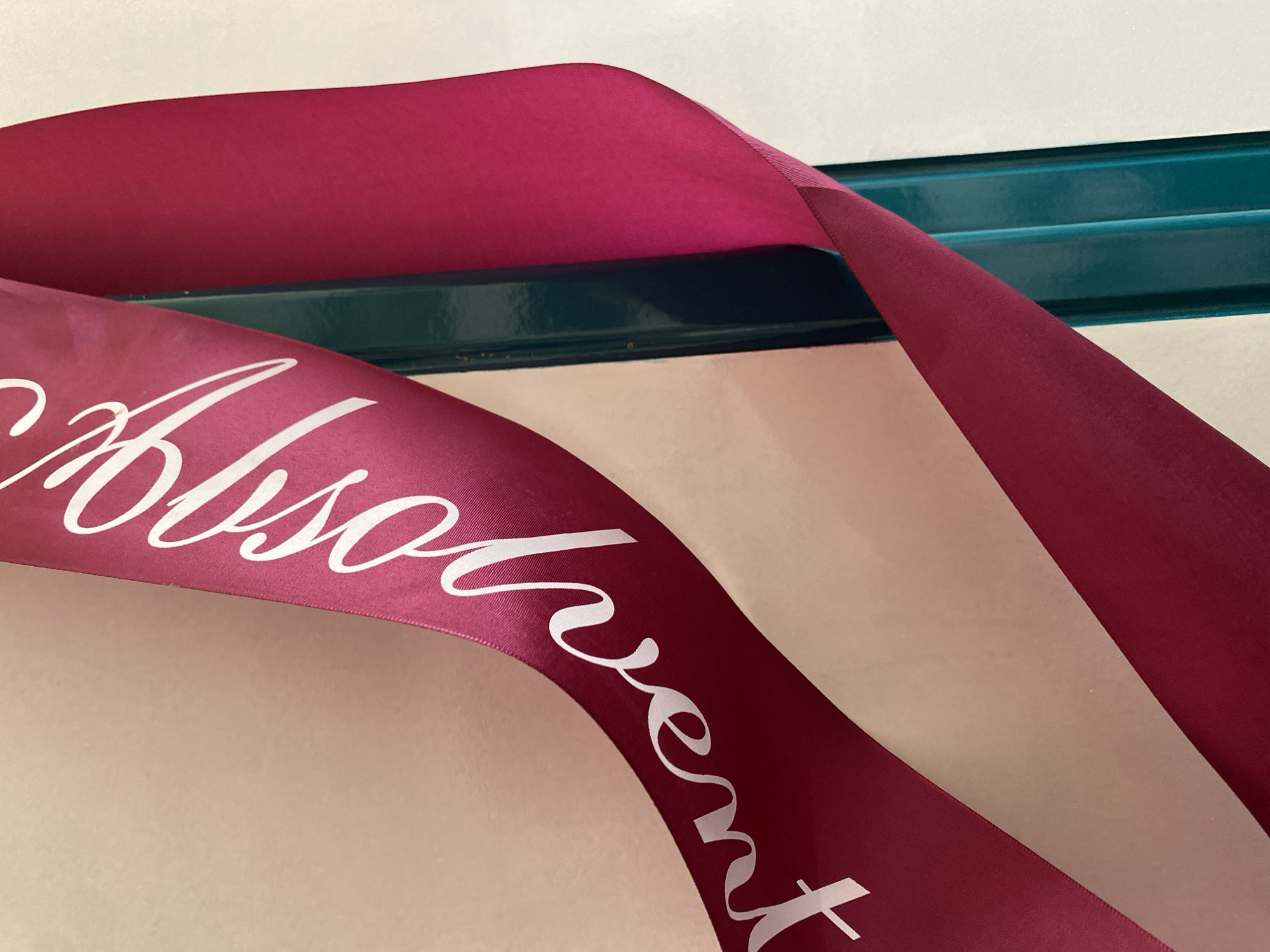 The image shows a close-up of a magenta ribbon with white text that appears to say "Březová". The ribbon is draped over a dark teal metal bar. The background is a textured, light-colored surface. The lighting suggests an indoor setting. There is no information about Bílina, Czechia in the image.