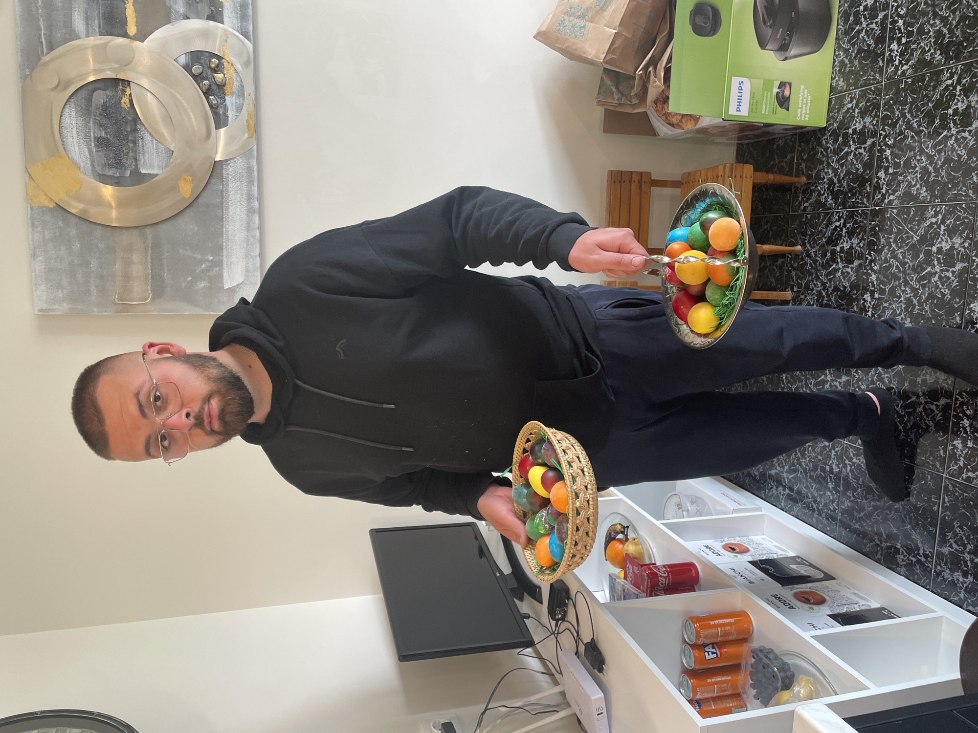 Here's a detailed description of the image:
**People:** A man with short hair, a beard, and glasses is the primary subject. He is wearing a black hoodie and black pants.
**Objects:**
* Two containers with colorful Easter eggs. One appears to be a woven basket, and the other is a metal bowl.
* A Philips All-in-One Cooker box.
* Wooden stools.
* A white shelving unit filled with items such as: cans, a small Coca-Cola box, boxes, glass bowls with food items, and electronic devices.
* A computer monitor.
* Wall art with concentric circles.
**Actions:** The man is holding out both containers of Easter eggs.
**Environment:** The setting is indoors, likely a kitchen or dining area. The floor is covered in a dark patterned tile. There's a white wall and a white shelving unit against the wall.
**Location:** While the image doesn't provide explicit information about the city of Sofia, Bulgaria, it is impossible to determine the location based solely on the visual elements.