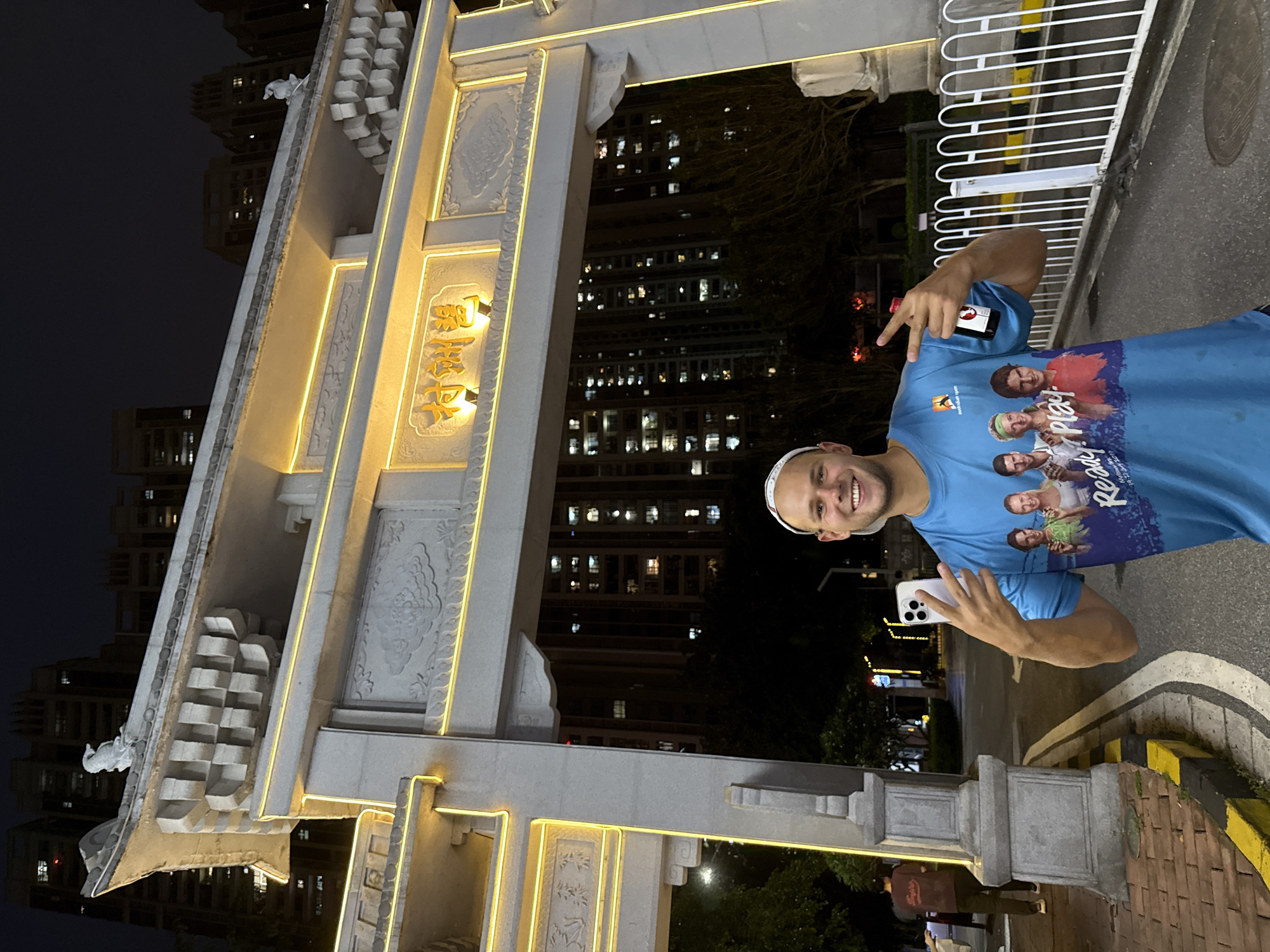 An outdoor nighttime scene depicts a Caucasian male, smiling broadly, positioned centrally in the foreground. He wears a blue t-shirt featuring images of tennis players and the text "Ready? Play! Australian Open," a white baseball cap turned backward, and dark shorts. His left hand holds a white smartphone with multiple camera lenses, while his right hand forms a peace sign and holds a small, dark-colored bottle with a red cap. The bottle's label is obscured.
Behind the man is a prominent traditional Chinese-style stone archway, illuminated with yellow LED strip lights outlining its structure and decorative elements. The archway features carved details, including cloud patterns and a small, white, bird-like statue on its sloped roofline. Centered above the main arch, illuminated in yellow, are three Chinese characters: "鹭洲村" (Lùzhōucūn), which translates to Luzhou Village. This location is situated in the Haizhu District of Guangzhou, China.
In the mid-ground, visible through and around the archway, are numerous multi-story residential buildings, their windows glowing with interior lights against the dark night sky. Trees and foliage line the street. To the left of the archway, a paved road with white and yellow markings is visible, alongside a brick sidewalk. Another individual, seen from behind, walks away on the left side of the road. A white metal fence is present on the right side of the frame. The overall impression is an urban or suburban setting at night.