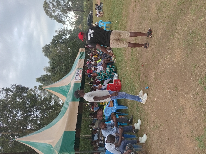 The image depicts an outdoor gathering in Chevaywa ward, Kenya. The scene features a group of individuals assembled on a grassy field under an overcast sky, with trees visible in the background.
Two large, green and beige striped tents are erected, providing shelter for the main crowd. A banner, partially visible on one tent, displays text including "WETU".
In the foreground, two adult males stand on a dirt patch surrounded by grass. The male on the left wears a light-colored t-shirt, dark baseball cap, blue jeans, and white sneakers, standing with his left side partially facing the viewer. The male on the right, with his back to the viewer, wears a black t-shirt with white text "MTETEZ WETU", a red baseball cap, khaki shorts, and dark sandals. A small blue object lies on the ground between these two individuals.
Beneath the tents, a large group of people, primarily adults, are seated on blue plastic chairs, facing towards the front of the gathering. Some red crates are visible near the seated attendees. To the far right, in the background, a dark vehicle is parked, and a smaller group of individuals are gathered around it on the grass. The overall scene suggests an organized outdoor community event or meeting.
