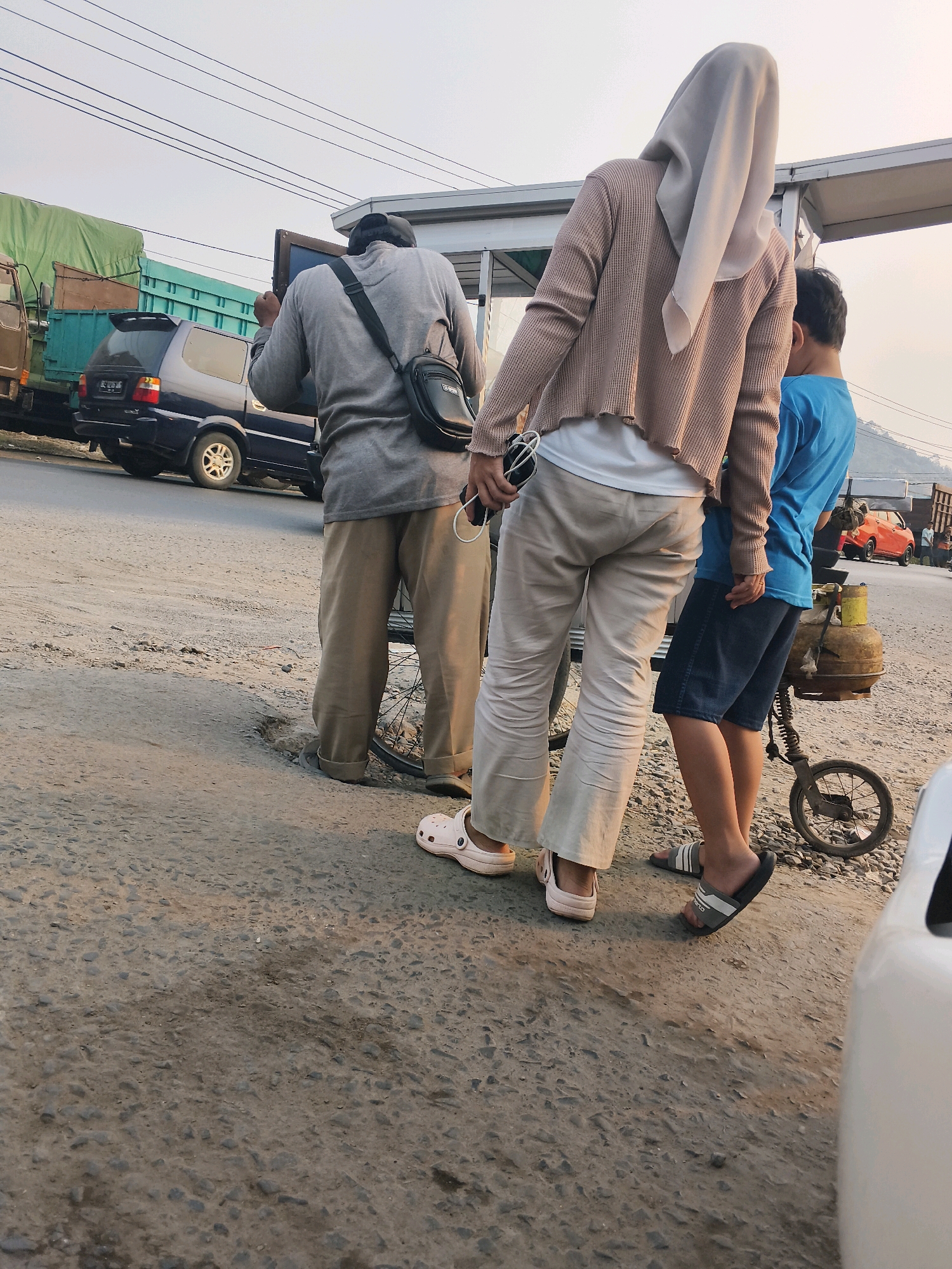 In Sabah Balau, Indonesia, a person wearing a grey long-sleeved shirt and khaki pants, with a black bag slung across their shoulder, stands on a dirt road. They are holding a flat-screen television. To their right, a person in a beige cardigan and light-colored pants, wearing a hijab, is also present, and a young person in a blue t-shirt and dark shorts stands next to them. A gas tank on a wheeled cart is visible behind the young person. In the background, trucks, a dark-colored SUV, and a small orange car are parked on the side of the road. The environment appears to be an outdoor market or roadside area with a clear sky overhead.