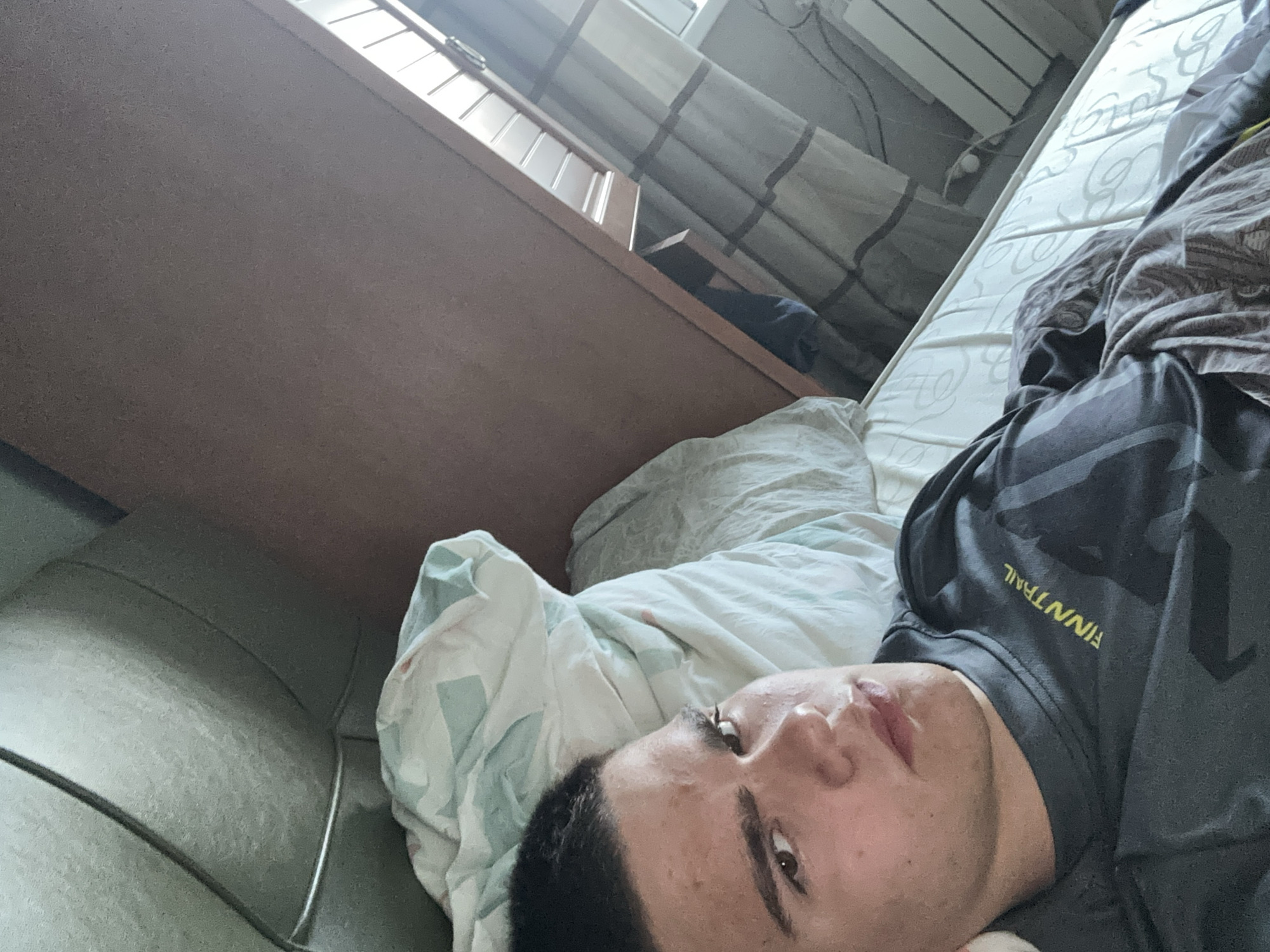 A person lies on a bed in a room. The person is wearing a gray t-shirt with "FINNTRAIL" printed in yellow. A bed with white patterned sheets and a duvet is beneath the person. A brown dresser is to the left of the bed. A window with sheer curtains is behind the dresser. The exact location is unknown.