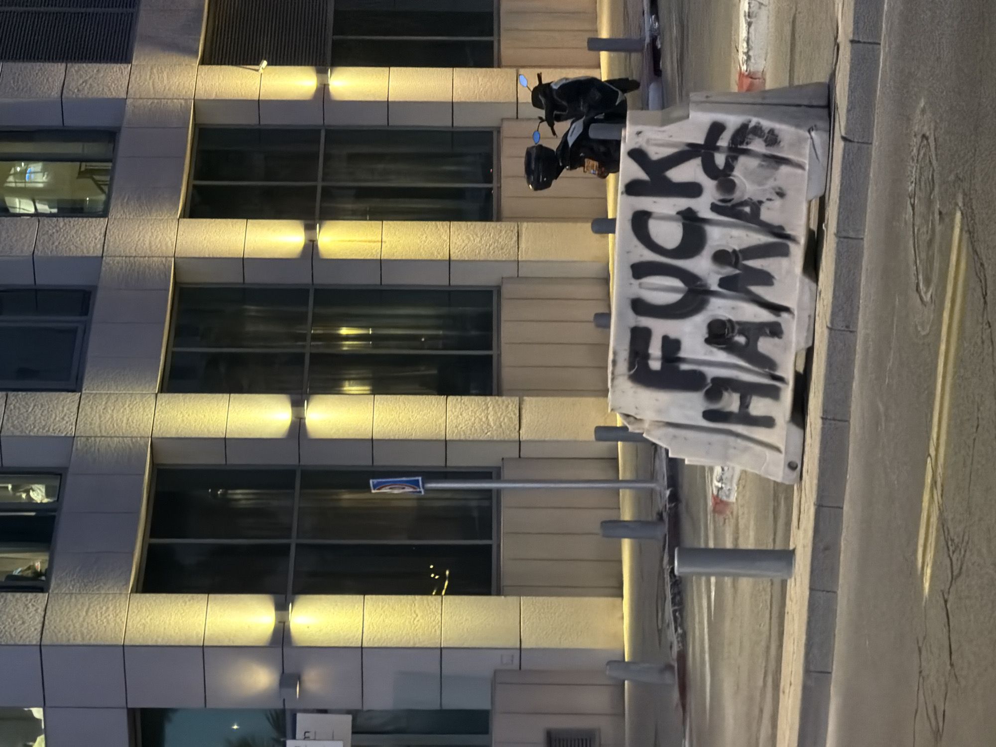 The image shows a low-angle view of a building facade with multiple windows, each having a row of horizontal lights above it. In front of the building, a white concrete barrier with "FUCK HAMAS" spray-painted in black is visible. A parked motorcycle is behind the barrier. A grey sidewalk runs alongside the barrier. The location is Tel Aviv, Israel.