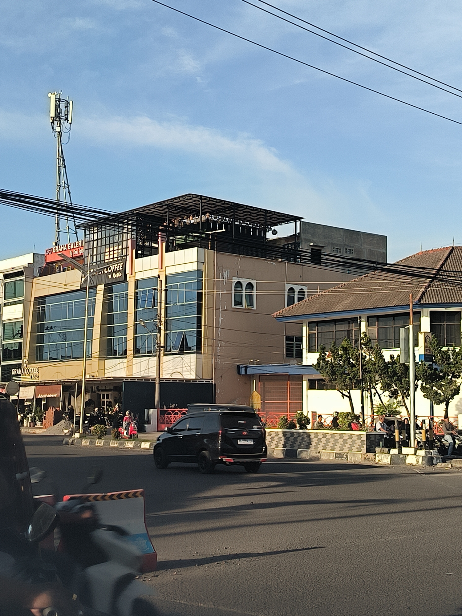 The image shows a street scene in Lhokseumawe, Indonesia. The street is paved and has moderate traffic, including a black SUV moving from left to right. A building with a "Graha Galeri Coffee" sign is visible on the left. A cellular tower rises above the building. Several pedestrians are visible on the sidewalks in front of the coffee shop and along the street. Another building with a tile roof is located to the right of the coffee shop. Power lines crisscross the sky. The sky is partly clear with a few clouds. A barrier, and parked motorcycles are also visible near the street. The weather appears to be sunny.