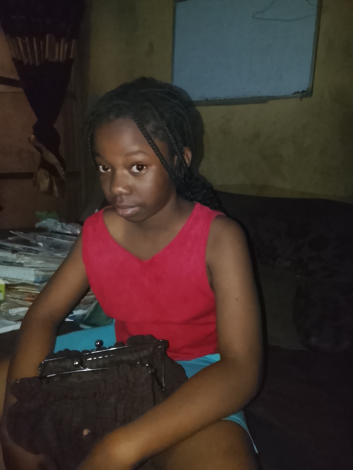 An indoor scene in Mgbidi, Nigeria, depicts a young female subject seated, facing forward, and looking directly towards the camera. The subject has dark skin and black braided hair. She is wearing a red sleeveless v-neck top and light blue shorts. Her hands rest in her lap, holding a dark brown, possibly fabric, handbag with a silver-colored metal clasp.

The background on the left side features a dark, draped fabric, possibly a curtain or garment, hanging vertically. Below this, a stack of papers or notebooks is visible on a surface. The background on the right consists of a light brown or beige wall with visible texture and discoloration. Mounted on this wall is a light blue rectangular object, possibly a board or frame, with a faint, light-colored drawing or mark near its top edge. A dark, possibly upholstered surface, which may be a bed or sofa, runs horizontally along the lower right portion of the background. The lighting appears dim, suggesting an internal light source or flash was used.