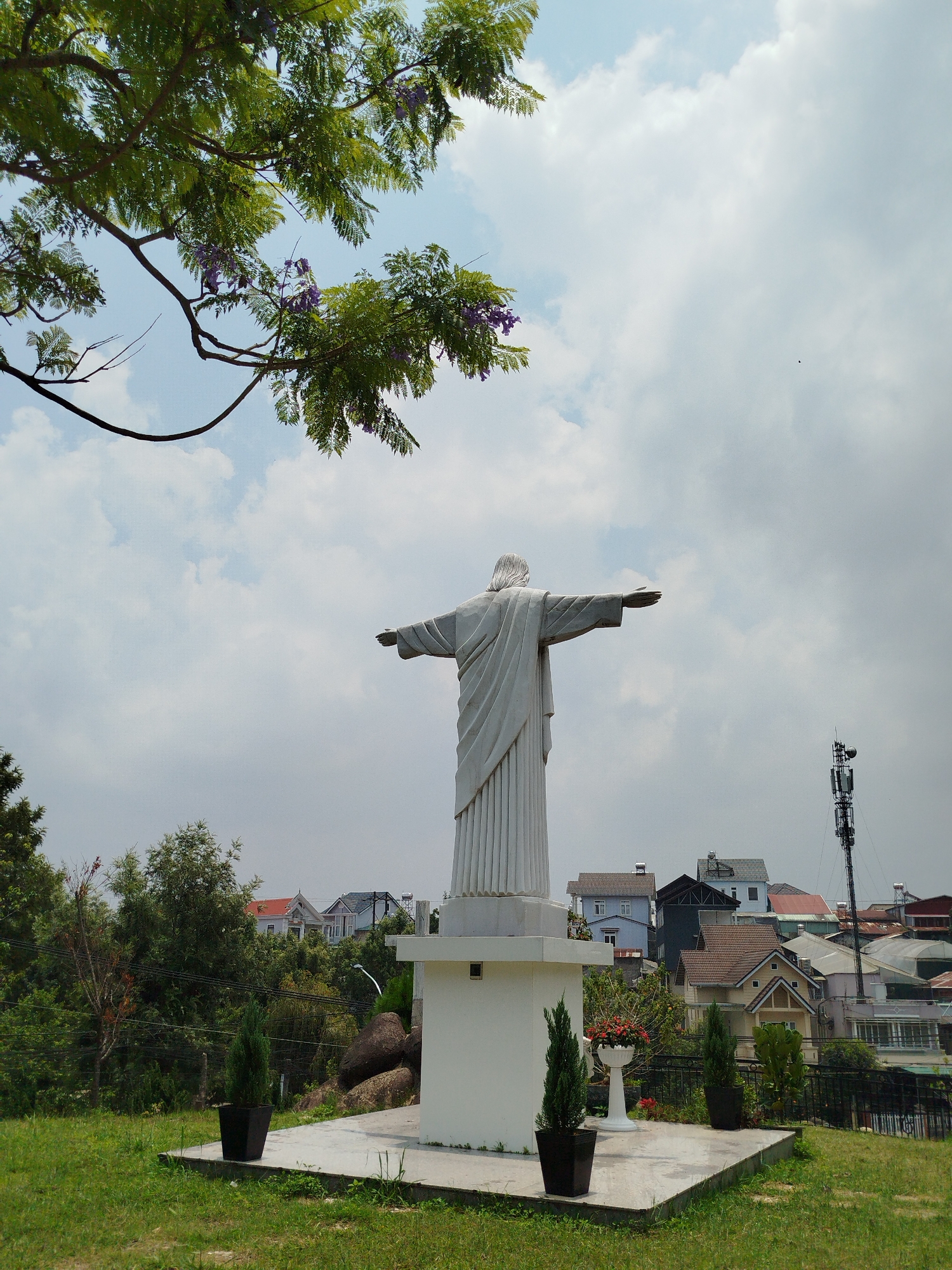 The image shows a white statue of Jesus Christ with arms outstretched, standing on a square white pedestal on a concrete base. The statue is positioned on a grassy lawn. In the background are trees, houses, and a telecommunications tower. A tree branch with purple flowers overhangs the statue from the top left corner. The sky is overcast with white clouds. The location is Lang Biang - Đà Lạt, Vietnam.
