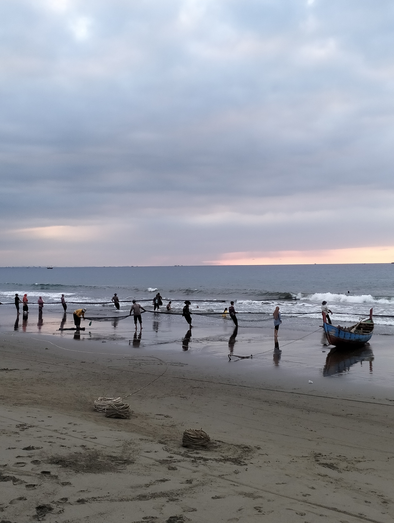 This image depicts a beach scene in Uleekareung, Indonesia, likely at dusk or dawn given the muted lighting and overcast sky. A group of approximately 15 individuals are engaged in pulling a fishing net from the sea towards the shore. Several people are in the water, while others are on the wet sand. One small, weathered blue and red fishing boat is partially submerged near the shore, with a rope attached to it, also being pulled by individuals on the beach. Several coiled ropes are visible on the sand. The ocean is calm with small waves, and distant boats are scattered on the horizon. The sky is a soft gradient of grey and pale pink, indicating diffused sunlight. The sand is wet and reflective, showing clear silhouettes of the people and the boat.