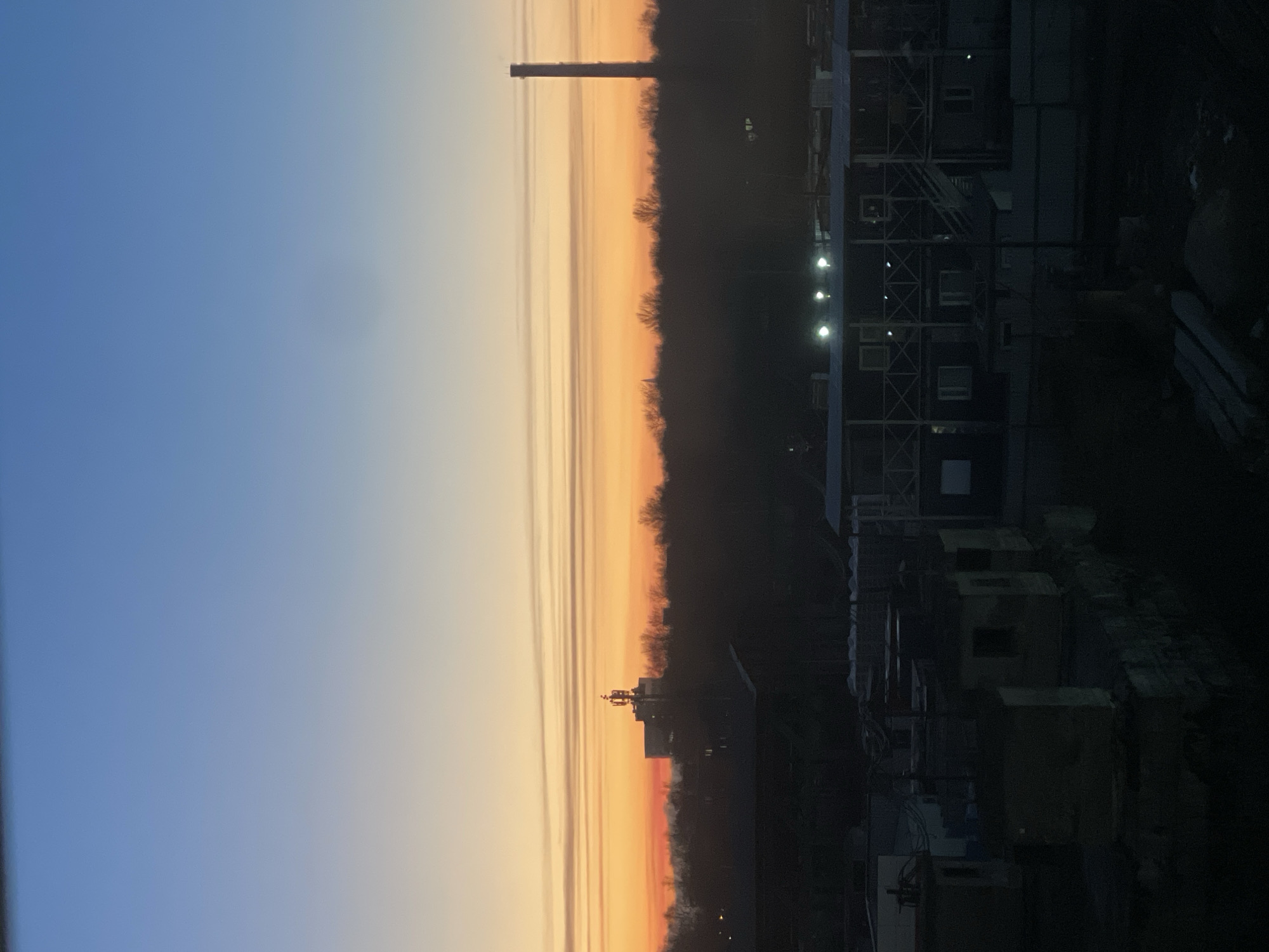 The image shows a sunset in Saint Petersburg, Russia, overlooking an industrial area. The sky transitions from blue to orange and red, with a silhouette of trees in the middle ground. A tall smokestack is visible on the left side of the skyline. Buildings and structures are visible in the foreground, some with lights on. The overall scene is dark.