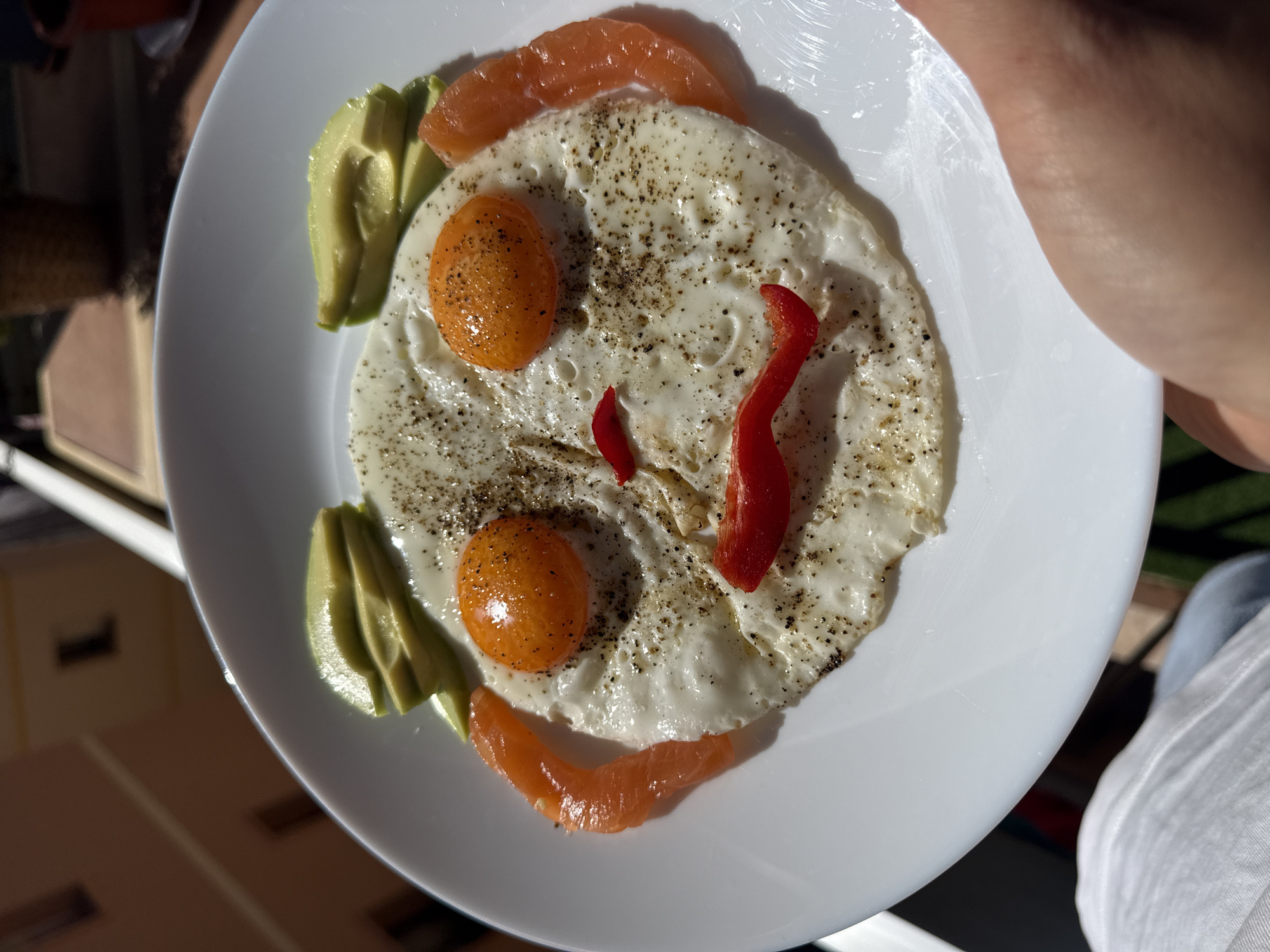 A top-down shot depicts a white plate held at an angle, presenting a stylized arrangement of food. Two fried eggs with bright orange yolks, heavily speckled with black pepper, form the "eyes" of a face. Slices of avocado are placed on the left side of the plate, while slices of salmon form the bottom portion of the face. Two pieces of red pepper are positioned as the mouth. The environment is out of focus and appears to be indoors.