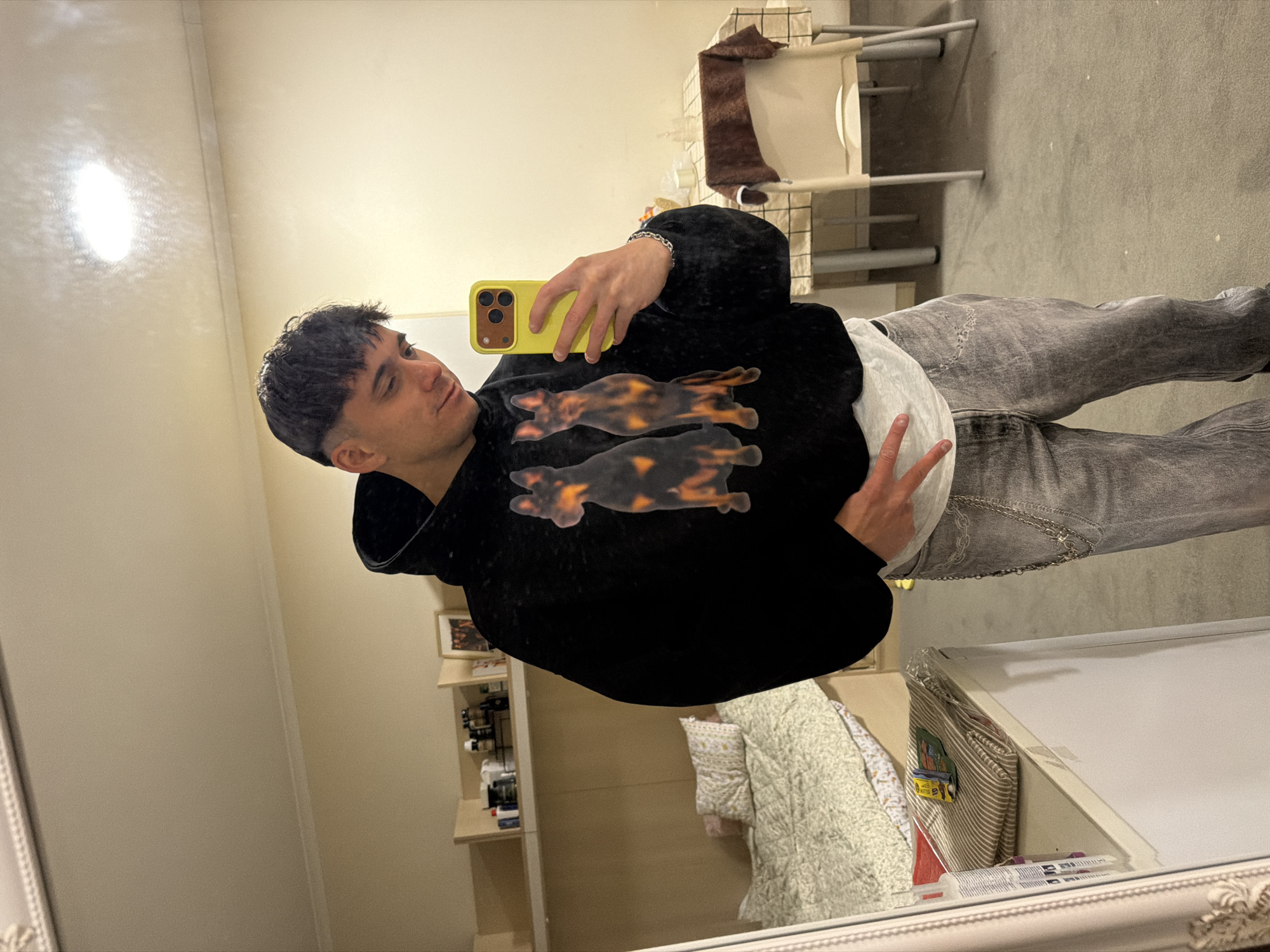 A young man is standing in what appears to be a bedroom or living space, taking a selfie in a mirror. He is wearing a black hoodie with a design featuring two dachshund-like figures on the front, light-colored jeans, and a white undershirt visible at the waist. He's holding a yellow smartphone in his right hand and making a peace sign with his left. The room contains a chair with a brown towel on it, a shelf with items, a bed with a quilted blanket, and a white surface with various objects. The environment suggests a residential space. The location within Sofia, Bulgaria, cannot be determined from the visual information provided.
