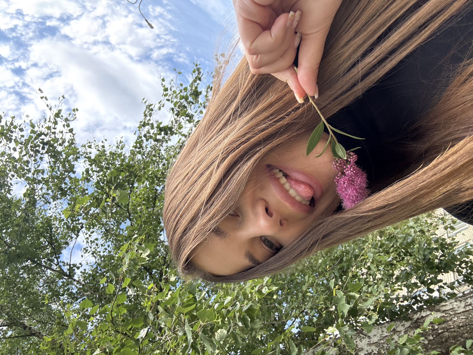 A young woman with long, brown hair is taking a selfie outdoors. She is wearing a black shirt. Her face is close to the camera, and she is sticking her tongue out. In her right hand, she holds a small green stem with pink flowers. The background consists of green trees and a blue sky with white clouds. The image appears to have been taken in the city of Azinsky, Russia, given the context provided.