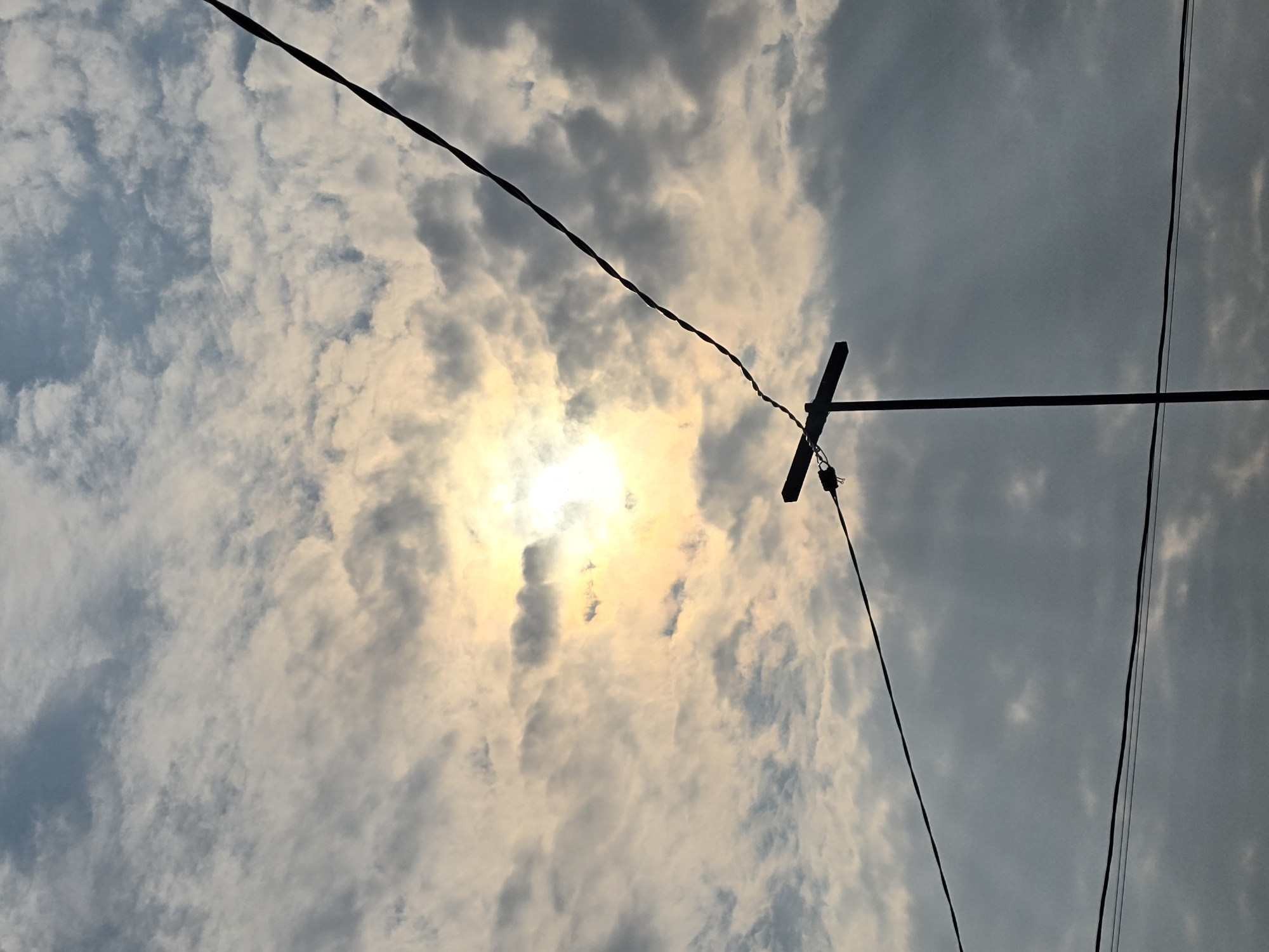 The image depicts a cloudy sky with the sun partially visible through the clouds, casting a yellowish glow. In the foreground, silhouetted against the sky, are utility poles and wires. One utility pole has a horizontal crossbar supporting wires, and another wire stretches diagonally across the frame from the lower left to the upper right. The scene is devoid of any people or discernable human activity. The image appears to be taken from a ground-level perspective, looking upwards.