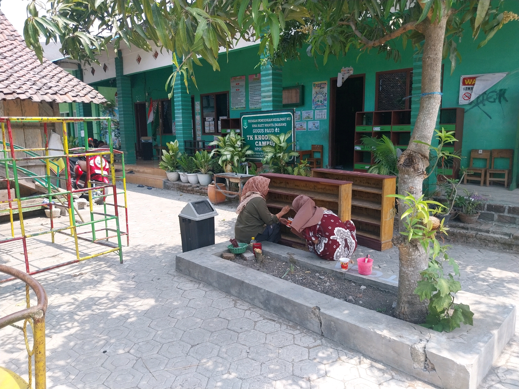 Two women are sitting on a paved courtyard outside a green building in the city of Campur, Indonesia. The woman on the left is wearing a brown hijab and a green shirt, and she is crouched down near a planter box. The woman on the right is wearing a pink hijab and a maroon and white patterned dress. She is also crouched down near the planter box and appears to be working with some small objects. A red scooter is parked nearby, and a playground structure is visible in the background. The building has a tiled roof and several windows and doors. There are plants in pots around the building, and a trash can is next to the planter box. The ground is paved with hexagonal tiles, and shadows from trees are cast across the area.