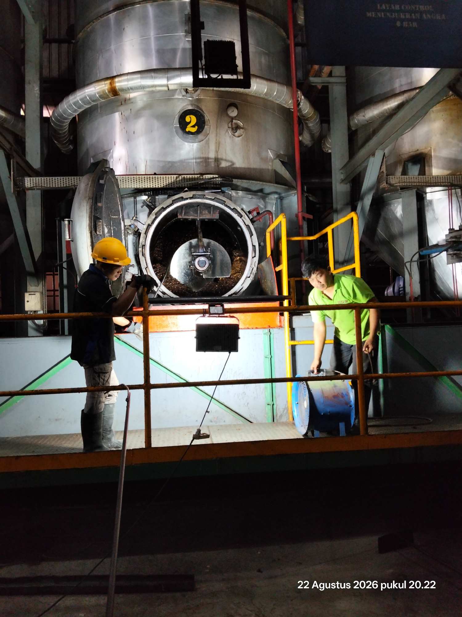 In an industrial setting in an unknown city in Indonesia, two men are visible on a raised platform next to large, cylindrical metal machinery. The man on the left, wearing a yellow hard hat, a dark shirt, and light-colored pants with black boots, stands leaning on a railing, looking down. He appears to be holding a tool. In front of him, the large machinery has an open, circular door revealing a dark interior filled with a granular substance.
To the right, a man in a bright green shirt and dark pants is interacting with a large, blue industrial fan. He is holding the fan by its handle and appears to be moving it. The environment is industrial, with metal structures, pipes, and railings. The lighting suggests it is nighttime or indoors with artificial lighting. A digital timestamp in the lower right corner reads "22 Agustus 2026 pukul 20.22".