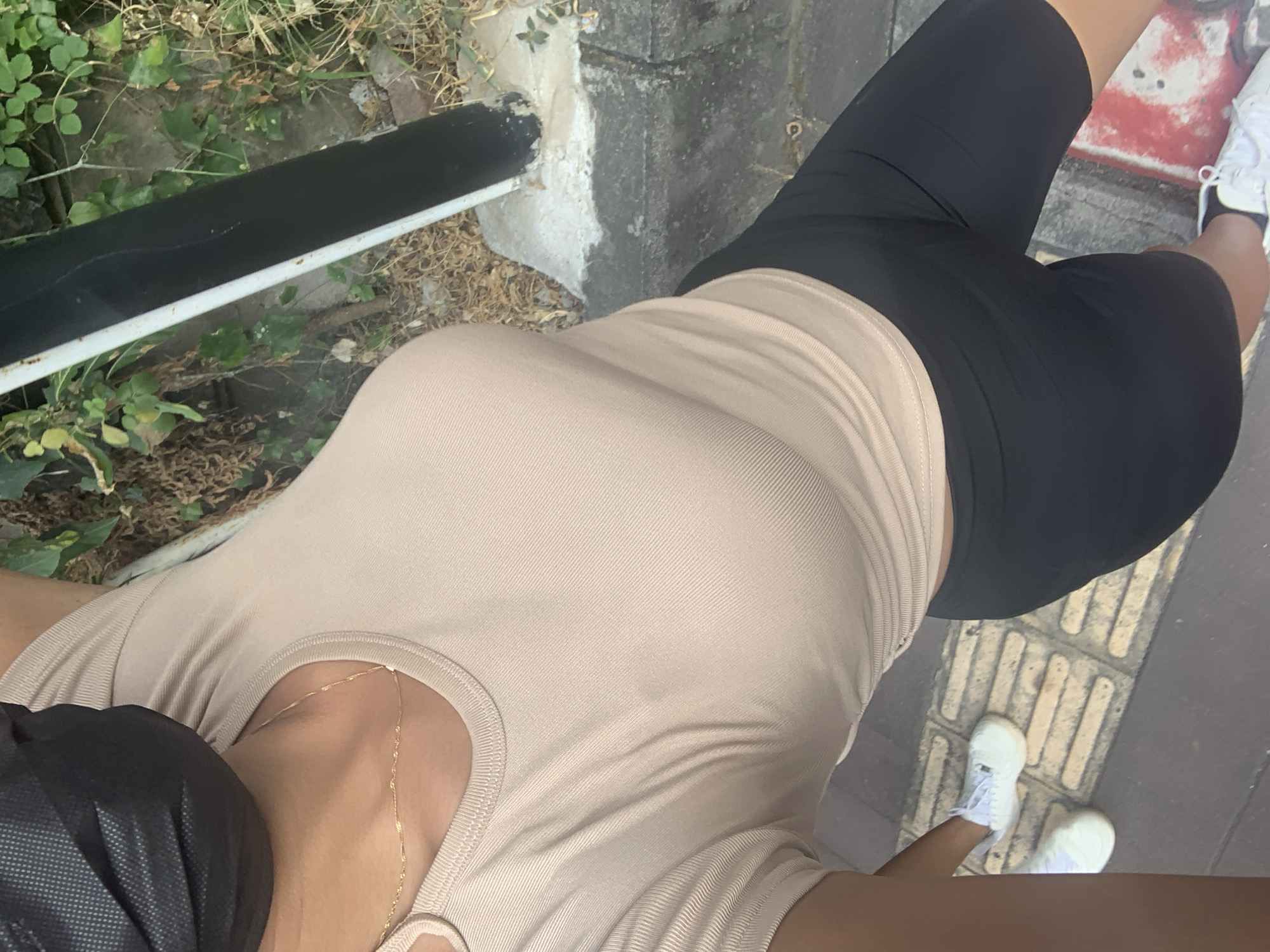 The image shows a close-up, top-down view of a person from the chest down, wearing a beige ribbed crop top, black bike shorts, and white sneakers. The person is also wearing a black face mask. They are standing on a textured sidewalk next to a grassy area with a black pole. The location is identified as Denpasar, Indonesia, though no specific landmarks or contextual clues within the image confirm this. The lighting suggests it is daytime.