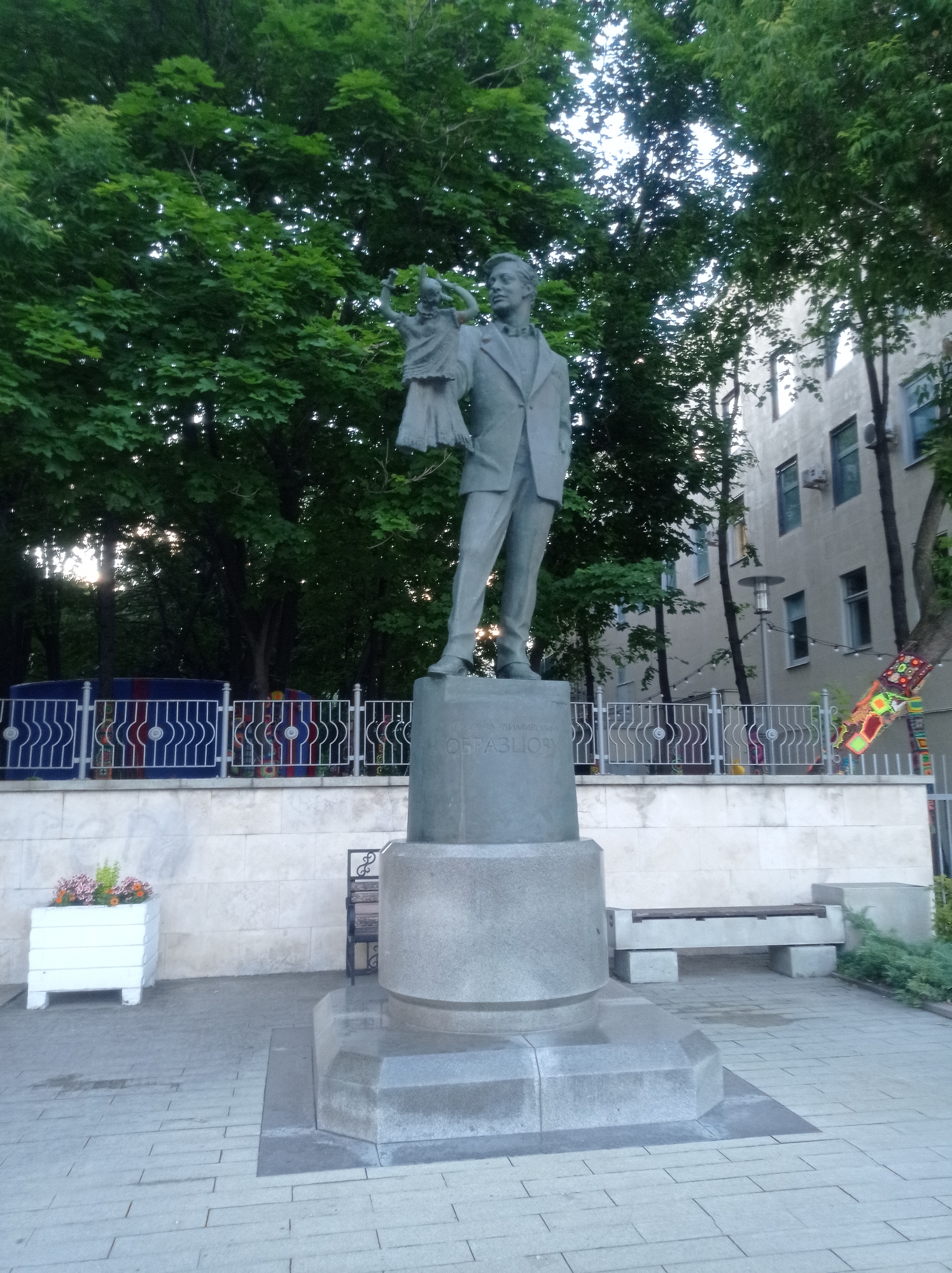 A statue of a man and child stands in an outdoor plaza. The man, dressed in a suit, is looking to his left and holding the child's hand. The child is reaching up towards the man. Behind the statue is a building with several windows. The plaza is paved with stones and there are trees and a fence in the background. The statue is made of bronze. The statue's inscription reads "ОБРАЗЦОВ". The statue is located in front of the Obraztsov Puppet Theatre in Moscow, Russia.