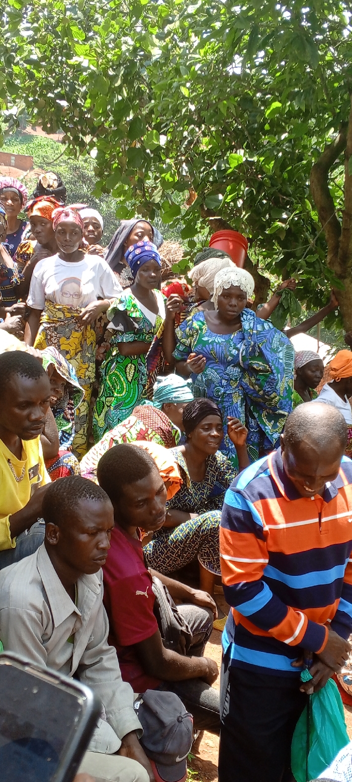 A diverse group of adults, mostly women, are gathered outside. They are dressed in various colorful fabrics and head coverings, with some wearing patterned dresses and scarves. Men are sitting in the front. A tree with green foliage partially obscures the background, hinting at a rural setting. One woman wears a white shirt with a portrait on it. A red bucket is visible behind the crowd. A mobile phone can be seen in the front of the image.