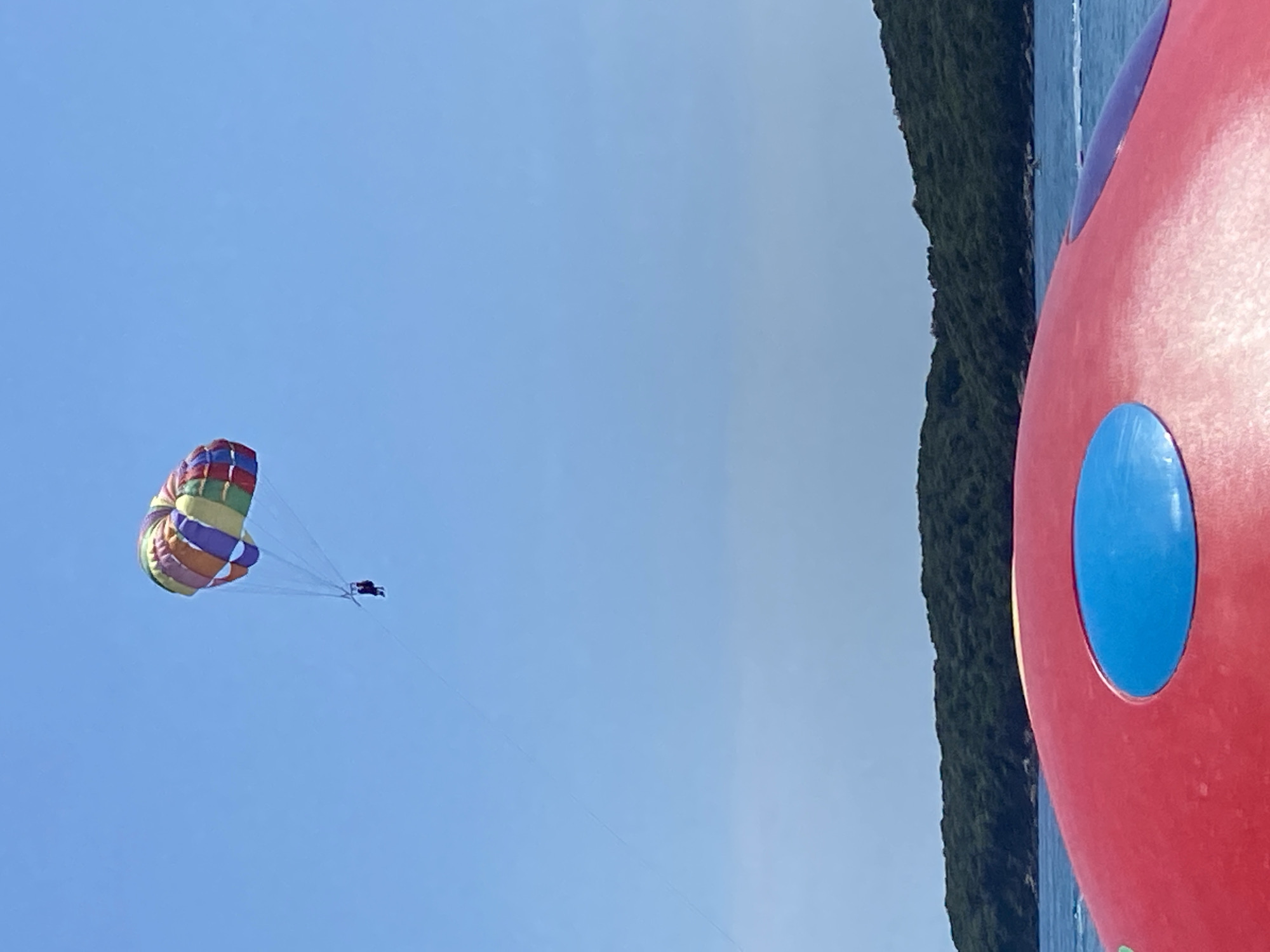 The image shows a parasailer in the blue sky in Prijevor, Montenegro. To the right is a small amount of trees and a part of a large red inflatable object with a blue circle on it. The water of the bay is visible.
