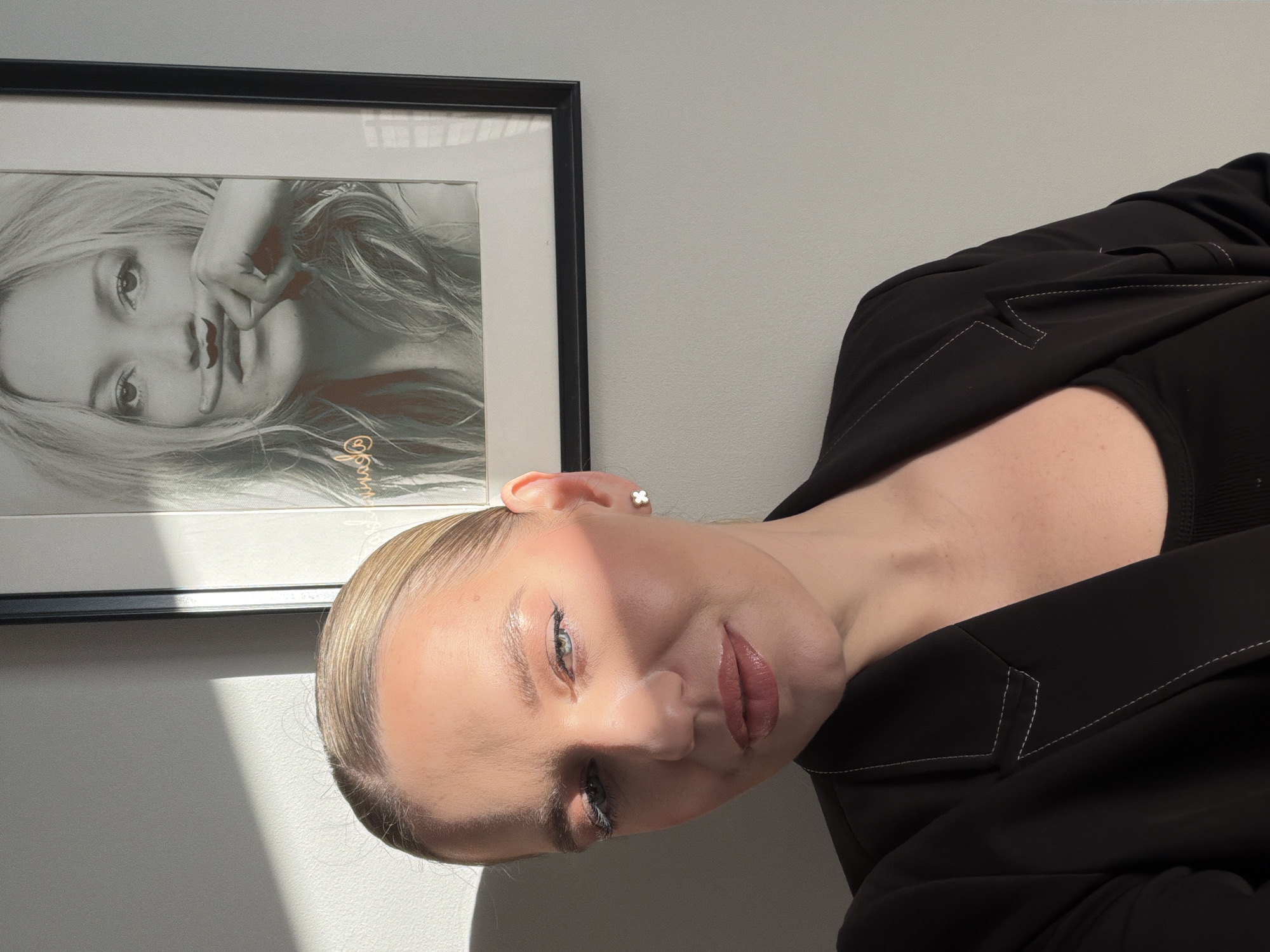 A fair-skinned woman with blonde hair is pictured indoors against a light gray wall. She wears a black top with a structured blazer with white stitching. Her hair is pulled back, revealing a small clover earring. The woman looks toward the camera. To her left, a framed black and white image of a woman is visible. The setting does not obviously indicate a specific location within Riga, Latvia. There is no readily available information about specific actions or interactions.