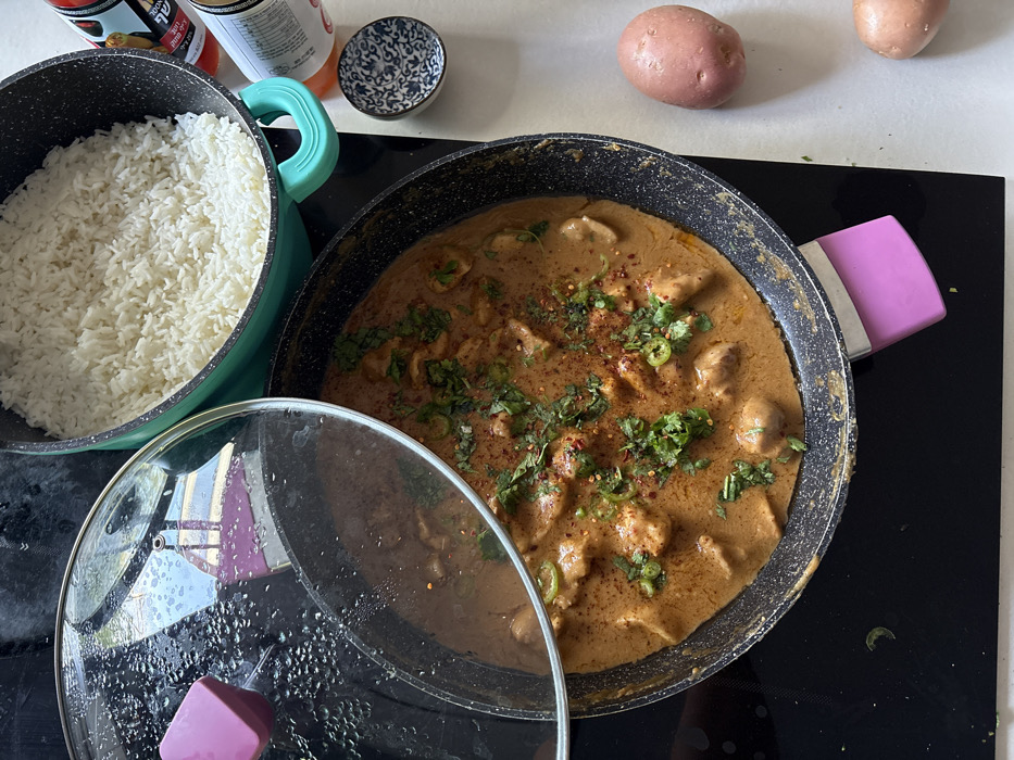 The image depicts a domestic kitchen scene in Raanana, Israel, showcasing a meal prepared on a black induction or ceramic cooktop and adjacent light-colored countertop.
In the foreground and center, a black non-stick pan contains a light orange-brown creamy stew, likely a curry, featuring multiple pieces of what appears to be chicken or other protein. The stew is garnished with chopped green herbs (consistent with cilantro/coriander), sliced green chilies, and sprinkled red pepper flakes. A clear glass lid, partially covered with condensation, rests askew on the pan, leaving a portion of the stew exposed. The pan is fitted with a pink silicone handle cover.
To the upper left, a teal-colored pot, also on the cooktop, is filled with cooked white rice. To the right of the cooktop, on the light-colored countertop, a small white bowl with blue patterned designs is visible. Behind this bowl, a partially visible bottle with an orange label displaying Hebrew text, including "שופרסל" (Shufersal, an Israeli supermarket chain), confirms the Israeli context. A white can or bottle is adjacent to it. To the lower right, two unpeeled, reddish-brown potatoes rest directly on the countertop. No individuals are present in the image.