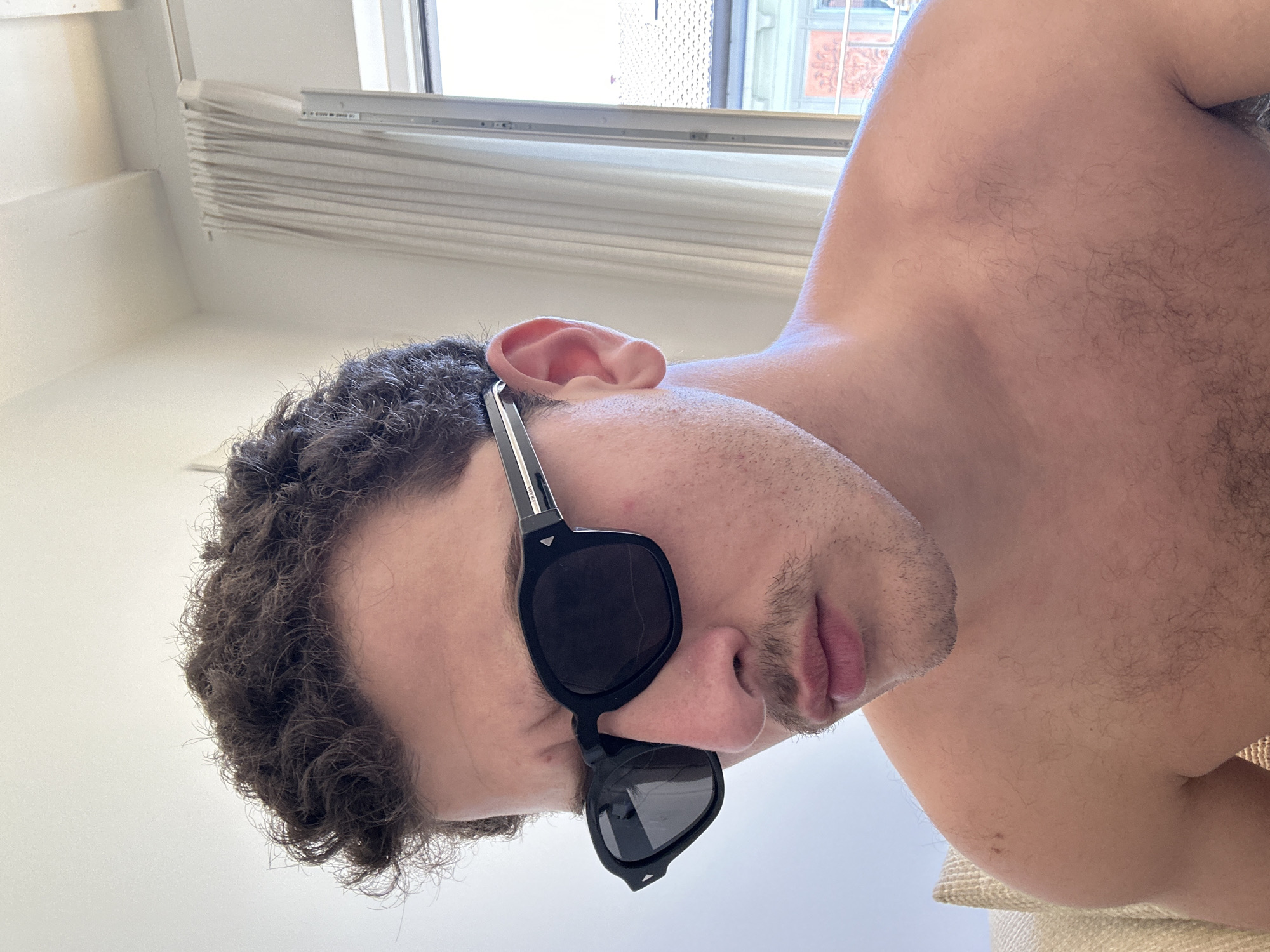 A low-angle, close-up shot of a shirtless man with curly, dark hair and stubble, wearing black sunglasses. The subject is likely inside an apartment, as indicated by the window blinds behind him. The window and buildings visible through the window suggest an urban environment that is possibly Milan, Italy. His face is partially obscured by the sunglasses. The room is bright and has white walls.
