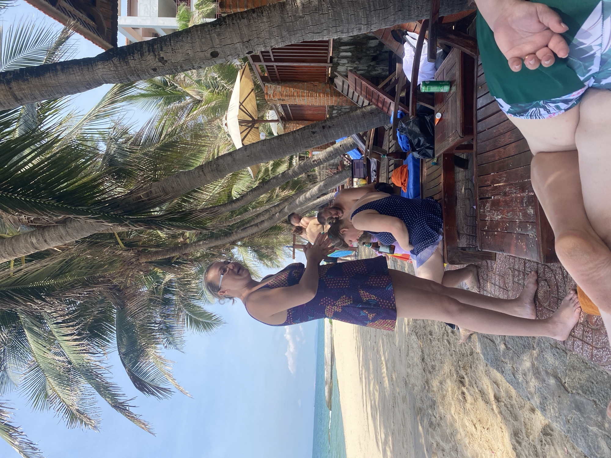 A group of people are relaxing on a beach in Phu Thuy Ward, Vietnam. The foreground shows the legs of a person wearing green patterned shorts. To the left, a woman in a purple patterned swimsuit stands and talks with another woman seated on a wooden bench. The seated woman is wearing a blue polka-dot swimsuit and has dark hair. Behind them, several wooden lounge chairs are arranged on the sand. A beach umbrella and a building with a red-tiled roof are visible further in the background. The scene is under a canopy of palm trees with the ocean and sky visible beyond.