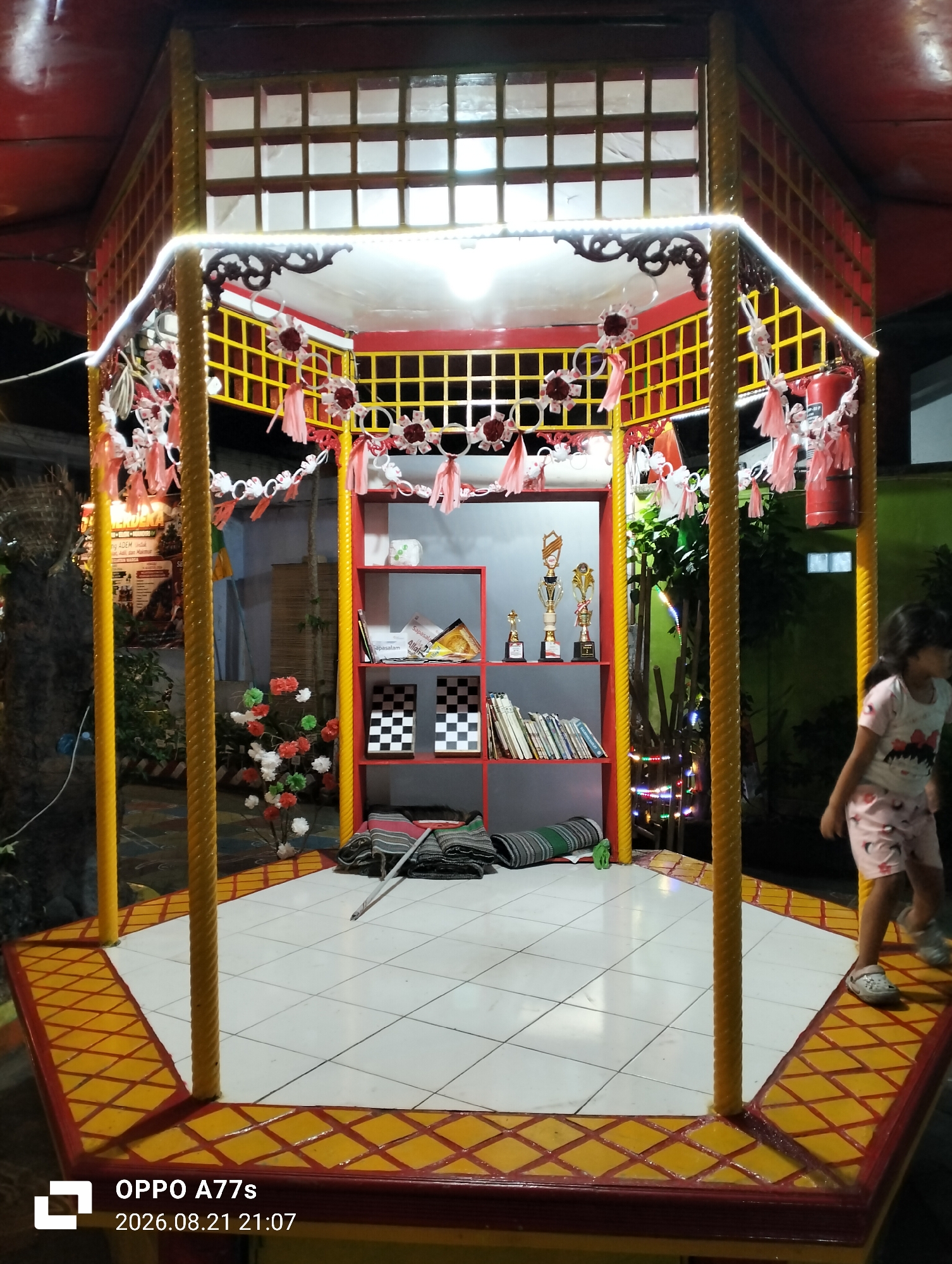 This image captures an outdoor gazebo with a decorative red and yellow roof. The gazebo features yellow rope-like pillars and is adorned with festive pink and white garlands and strings of lights. Inside, a red shelf unit displays several trophies and books. On the floor, there are rolled-up mats and some artificial flowers. A young girl in pink shorts and a t-shirt is visible walking on the right side of the frame. The foreground shows a tiled floor with a patterned border. The timestamp on the image indicates it was taken on August 21, 2026, at 21:07. The location is stated as Semarang, Indonesia.