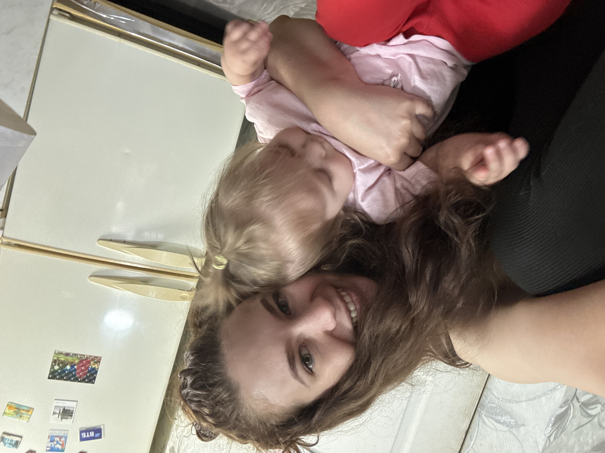 A woman with brown hair is smiling at the camera, with a young child resting on top of her. The child has blonde hair and is wearing a pink shirt. The woman is wearing a red shirt. They appear to be in a kitchen with a refrigerator visible in the background. The location is unknown.