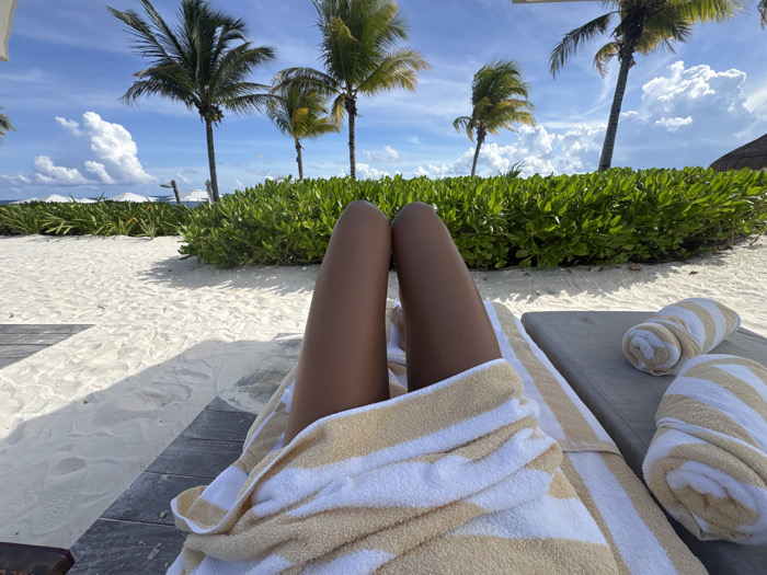 The image depicts a first-person view from a recumbent position on a beach in Marina Maroma, Mexico. The foreground features the upper legs of an individual, tanned, partially covered by a white and beige-striped towel draped over the lap. To the immediate right, a padded grey chaise lounge holds two additional, rolled-up towels with matching white and beige stripes. The surface beneath the chaise lounge and the visible parts of the legs is white sand, with a section of weathered wooden planks visible on the lower left.
Beyond the immediate foreground, a dense line of vibrant green foliage extends horizontally across the midground. Above this foliage, several tall palm trees with green fronds are visible against a bright blue sky dotted with prominent white cumulus clouds. In the distance, beyond the foliage, a body of water is discernible, with several white beach umbrellas scattered along what appears to be a shoreline. A segment of a thatched roof is partially visible on the far right. The scene is illuminated by natural sunlight, casting shadows on the sand.