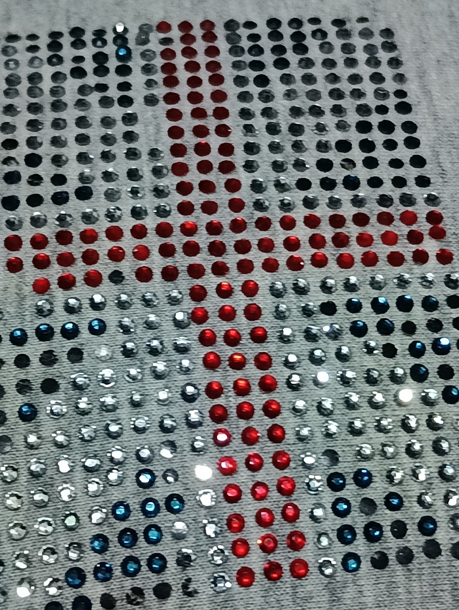 The image shows a close-up of a light-colored fabric, likely denim, adorned with rows of small, colorful rhinestones. These rhinestones are arranged in a grid pattern, forming a large design. The colors of the rhinestones are primarily red, white (represented by clear or silver rhinestones), and blue. The arrangement of these colors suggests a flag or a patriotic symbol. The fabric appears to be a piece of clothing or textile, possibly a shirt or a patch, given the way the rhinestones are densely applied. The lighting highlights the faceted nature of the rhinestones, causing some to sparkle brightly. The image is taken from a slightly elevated angle, looking down at the textured surface of the fabric and the embedded embellishments. There is no visible context of a specific location in Biliaivka, Ukraine, or any indication of people or actions within the frame.