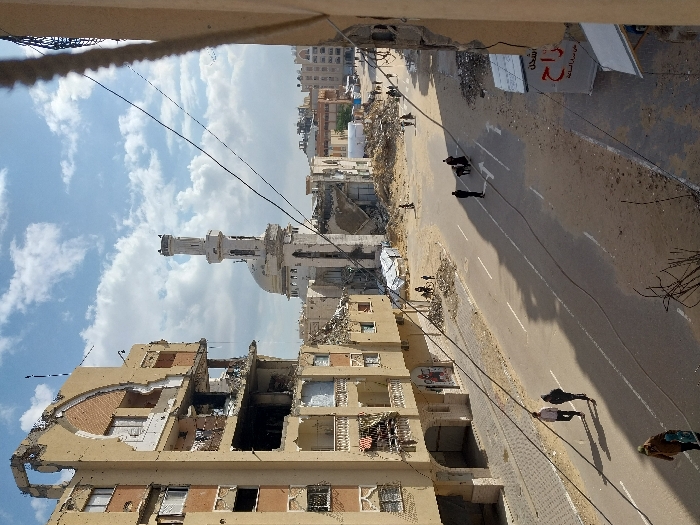 This image depicts a scene of extensive urban destruction in Khan Yunis, Palestinian Territory, viewed from an elevated perspective.
In the left foreground, a multi-story building, light brown/beige in color, stands severely damaged, with large sections of its facade and multiple balconies collapsed or missing, exposing interior rooms and structural elements. Broken windows and visible rebar are present.
A paved asphalt road, marked with white double solid and dashed lines, traverses the mid-ground. Patches of sand and debris are visible along its edges and partially cover the surface. Several overhead wires stretch across the frame, obscuring parts of the view.
On the road, two individuals walk side-by-side towards the lower left, casting long shadows. Further along the road, a small group of people are gathered or walking. In the far mid-ground, a white tent-like structure is visible amidst rubble.
Central to the mid-ground is a mosque, characterized by a large dome and a tall, slender minaret. The mosque itself exhibits significant structural damage, with visible breaches in its walls and roof. The area surrounding the mosque is completely reduced to rubble and debris.
To the far right, a partial view of another damaged building with rough edges and exposed interior is visible. A white sign with red Arabic script is attached to this structure.
The background consists of a sprawling landscape of heavily damaged or destroyed buildings and mounds of rubble, indicating widespread devastation. The sky is partly cloudy with patches of blue. The scene is illuminated by daylight, casting distinct shadows from the structures and individuals.