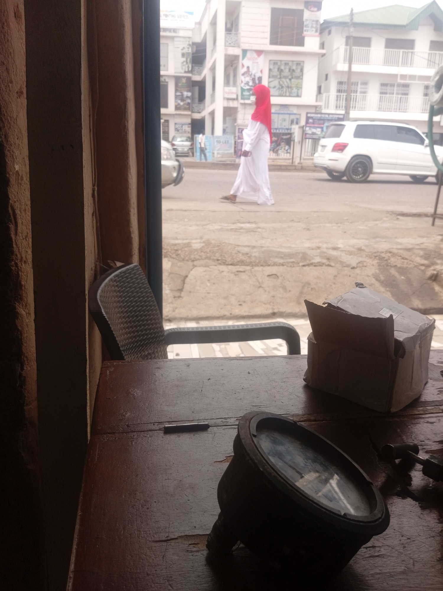 This is a medium shot of a street scene captured from an interior, likely through a window. In the foreground, on a wooden table, rests a black, circular object with a glass face, possibly a gauge or a clock, and a damaged cardboard box. To the left, a portion of a dark-colored chair with a woven seat is visible.
The background depicts a street with a white SUV parked on the right. A person wearing a white abaya and a red hijab walks across the street, facing left. Buildings with multiple stories line the street, featuring white facades and balconies. Some of these buildings have signs, suggesting commercial establishments. The street appears to be paved, with some visible cracks and wear. The sky is overcast. The scene is consistent with a location in Sardauna, Nigeria, where such architecture and street life are common.