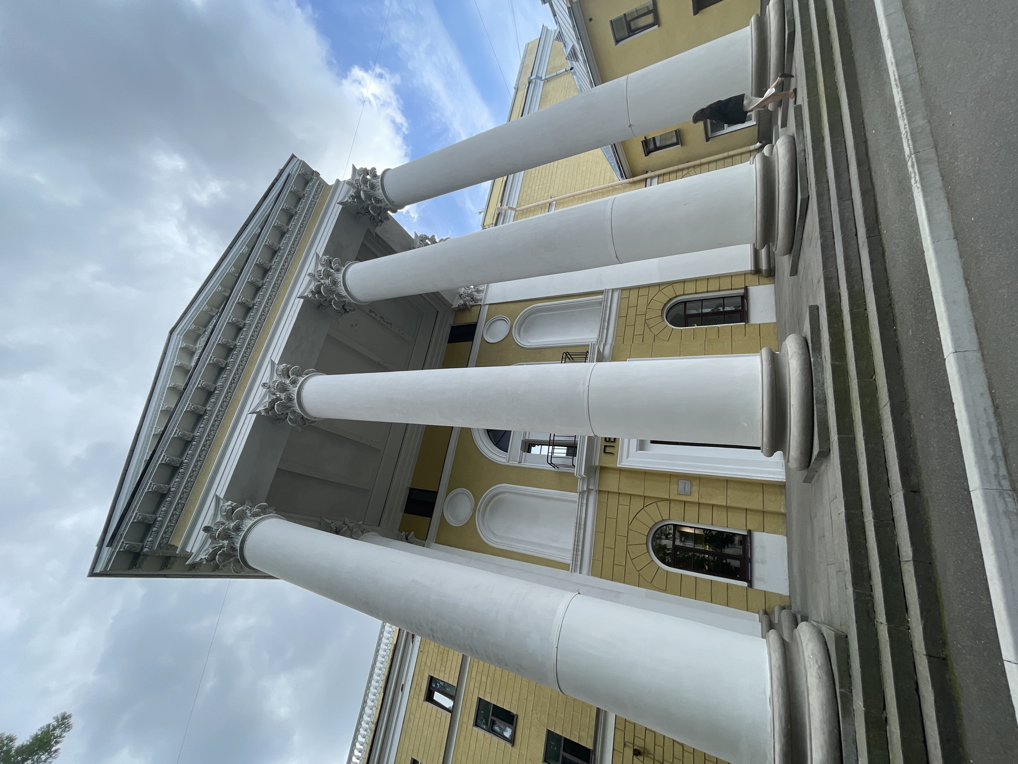 This media file depicts a low-angle shot of a yellow building with classical architecture, characterized by large white columns and ornate detailing on the facade. The building appears to be a grand structure, possibly a museum, government building, or historical landmark. Sunlight illuminates the building, casting shadows and highlighting the architectural features, suggesting it is daytime. The sky is partially cloudy, indicating overcast weather.
A person is visible on a ledge near the top of the building, adding a human element to the scene and providing a sense of scale. The person's posture and presence might suggest contemplation or a moment of pause. The image is oriented with the building's facade running diagonally across the frame, creating a dynamic perspective.
The scene is set in Saint Petersburg, Russia, a city known for its historical and architectural significance. While no specific text is clearly legible in the image, the style of the building is consistent with the neoclassical architecture found in the city. The overall impression is one of grandeur and history, with a touch of human presence within the imposing structure.