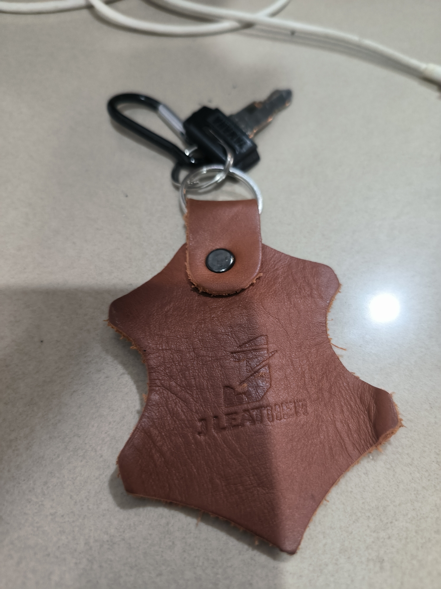 The image presents a close-up view of a keychain, featuring a brown leather tag, a metal key, and a black carabiner clip, all resting on a light-colored, possibly tiled or laminated, surface. The scene suggests an indoor setting, likely on a table or countertop.
The central object is an irregularly shaped leather tag, resembling a piece of hide, with a noticeable grain texture. A metal rivet or snap button secures a small leather loop at the top of the tag. Connected to this loop via a silver split ring are a black carabiner clip and a single silver-colored key with a dark, possibly plastic, key head.
Prominently embossed on the leather tag is the text "J LEATHER", positioned beneath a small, stylized logo that is somewhat indistinct but appears to be part of the brand's emblem. A bright light reflection is visible on the surface to the right of the leather tag. In the blurred background, a white cable or cord is present. The overall image depicts these objects in a static arrangement, with no discernible activity taking place. The media file was taken in Lhokseumawe, Indonesia.