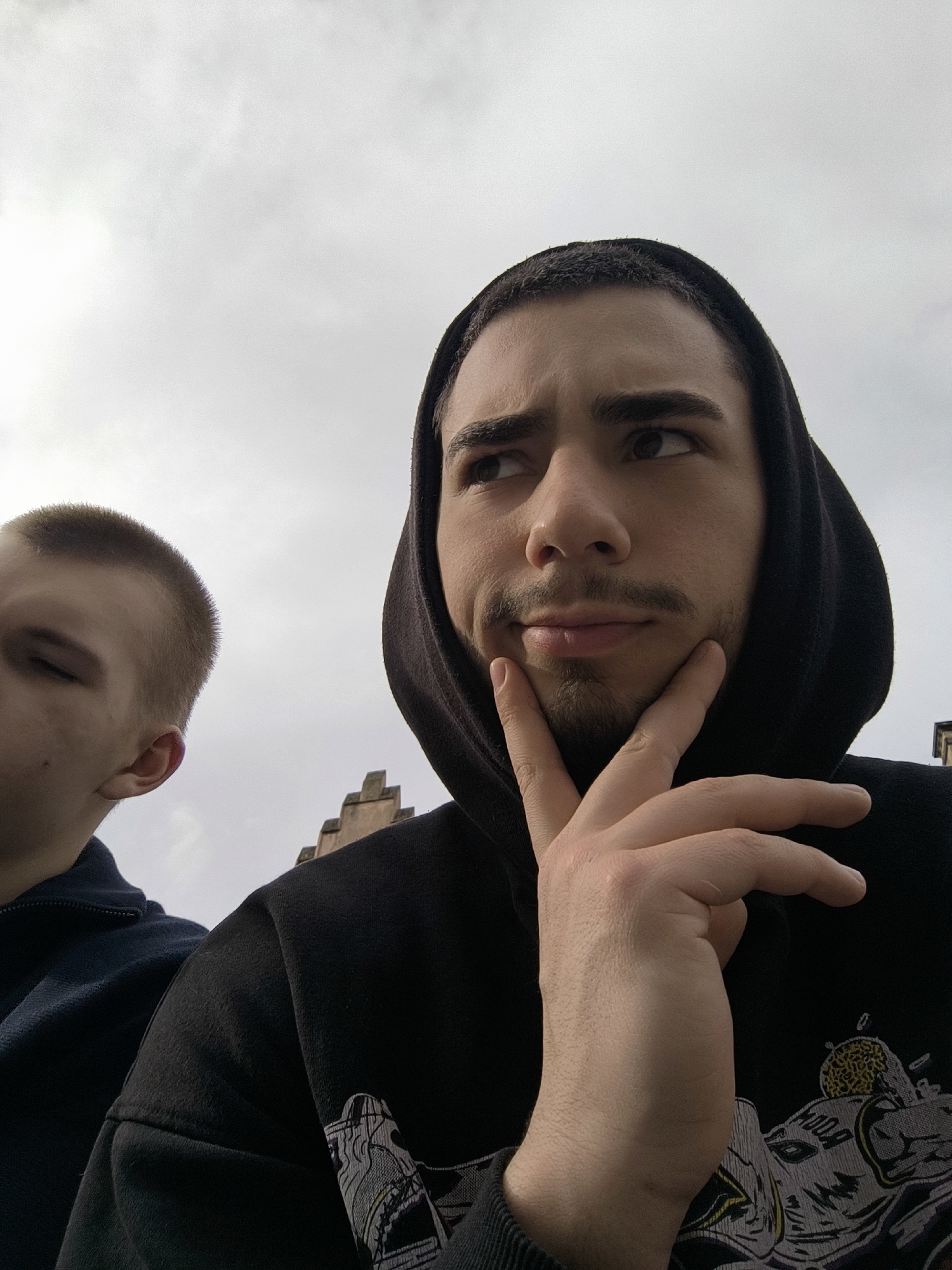 Two young men are standing outside in Strasbourg, France. The man on the right, closest to the camera, is wearing a black hoodie and has dark hair and a goatee. He is resting his hand on his chin and looking upwards with a thoughtful expression. The other man, partially visible on the left, has a buzz cut and is wearing a dark blue jacket. In the background, there is a building with a distinctive stepped gable roof, a common architectural feature in Strasbourg. The sky above is overcast.