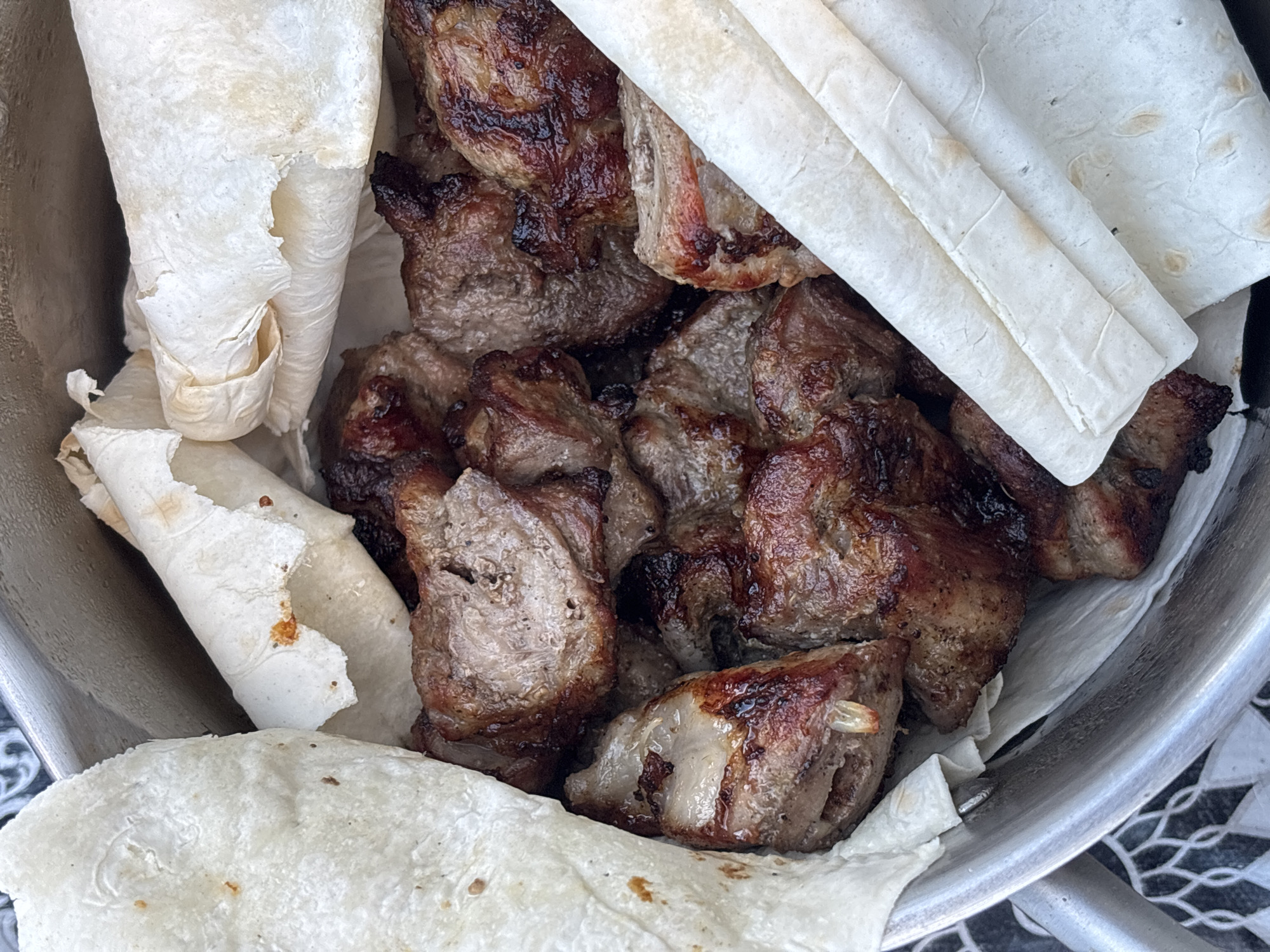A close-up, overhead shot shows a metal bowl filled with grilled meat skewers and pieces of flatbread. The meat is a dark brown, with some charred edges, and appears to be tender and juicy. The flatbread is thin and white, with some slightly browned spots, and is folded and tucked around the meat. The background is blurred, but suggests an outdoor setting, possibly a picnic or barbecue. There is a patterned surface visible at the bottom of the frame. The image was taken in the city of Мещёры, Russia.
