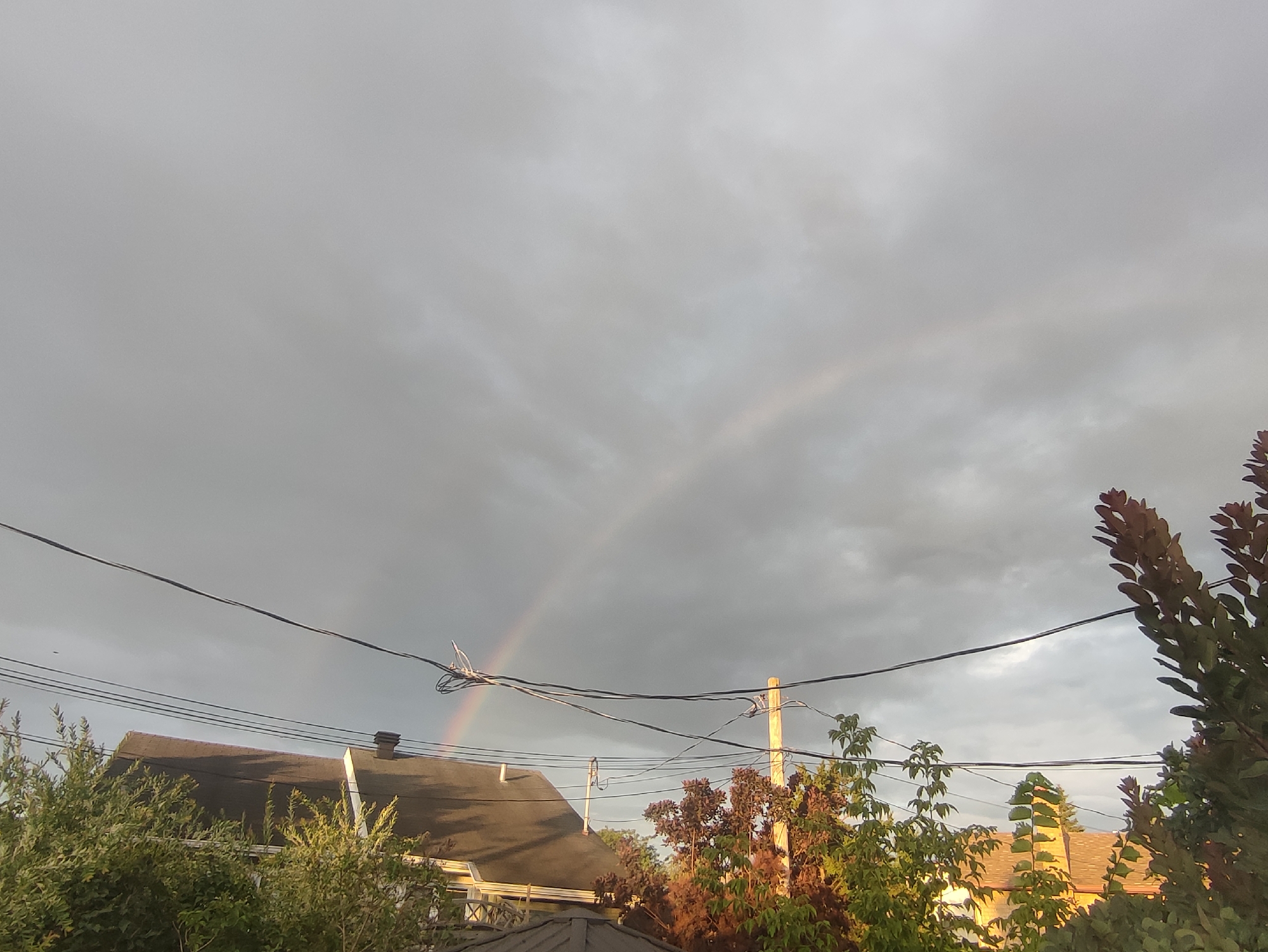 The image shows a suburban street scene in Longueuil, Canada, under a cloudy sky with a faint rainbow visible. The foreground is dominated by lush green trees and some foliage with reddish-brown leaves. In the middle ground, the rooflines of several houses are visible, along with a prominent telephone pole and several overhead power lines crisscrossing the scene. The sky is predominantly gray with some lighter patches, suggesting recent rain or an approaching storm. The faint rainbow arches across the sky, partially obscured by the clouds. The overall impression is of a quiet residential area after a rain shower.