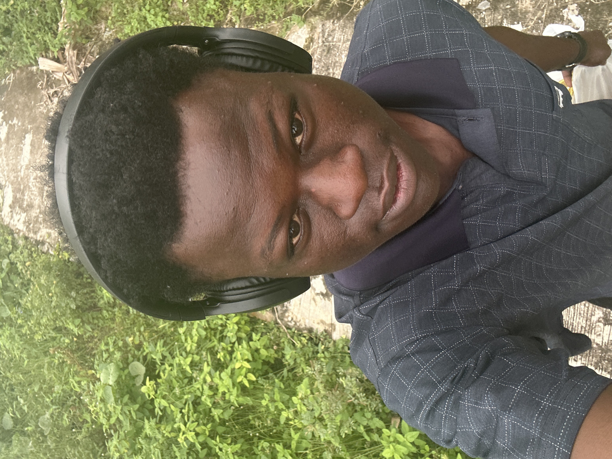 An outdoor, close-up, high-angle shot shows a young Black person wearing black headphones. They are positioned in the center of the frame, looking directly at the camera. The person has dark skin, short, curly black hair, and is wearing a dark blue collared shirt with a white checkered pattern. The background consists of lush green foliage and a textured, concrete pathway. The scene is likely located in Badagry, Nigeria, given the typical vegetation and the style of pathway.