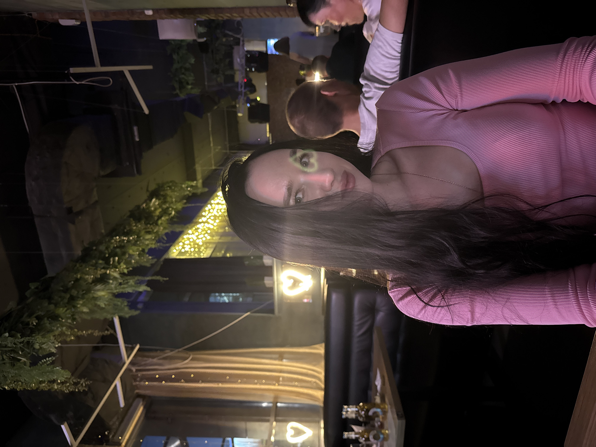The image depicts a woman with long, dark hair and a pink ribbed top, seated in what appears to be a dimly lit establishment. There is a green heart-shaped overlay on her left eye. The background suggests an indoor venue with seating, possibly a bar or restaurant. In the distance, other patrons are visible, and the decor includes hanging greenery and illuminated heart shapes. The overall ambiance is suggestive of a nightlife setting, possibly in Novosibirsk, Russia, given the context.