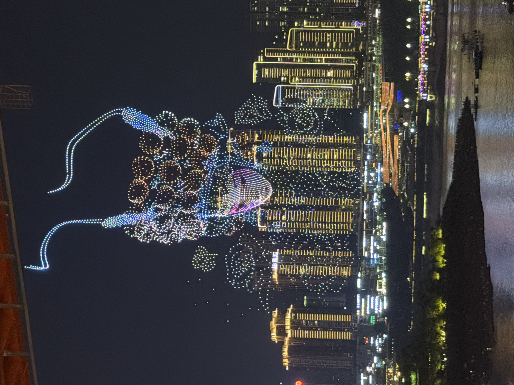 The image captures a nighttime drone show above the Yuzhong District in Chongqing, China. A large formation of illuminated drones in the sky depicts a stylized face wearing a mask and what appears to be hair or a headdress, rendered in a colorful mosaic of lights. Below the drone display, a cityscape of numerous high-rise buildings is illuminated with lights, primarily in yellow and white, creating a vibrant urban backdrop. A river flows in the foreground, with boats visible on its surface. The scene suggests a festive or celebratory event taking place in the city.