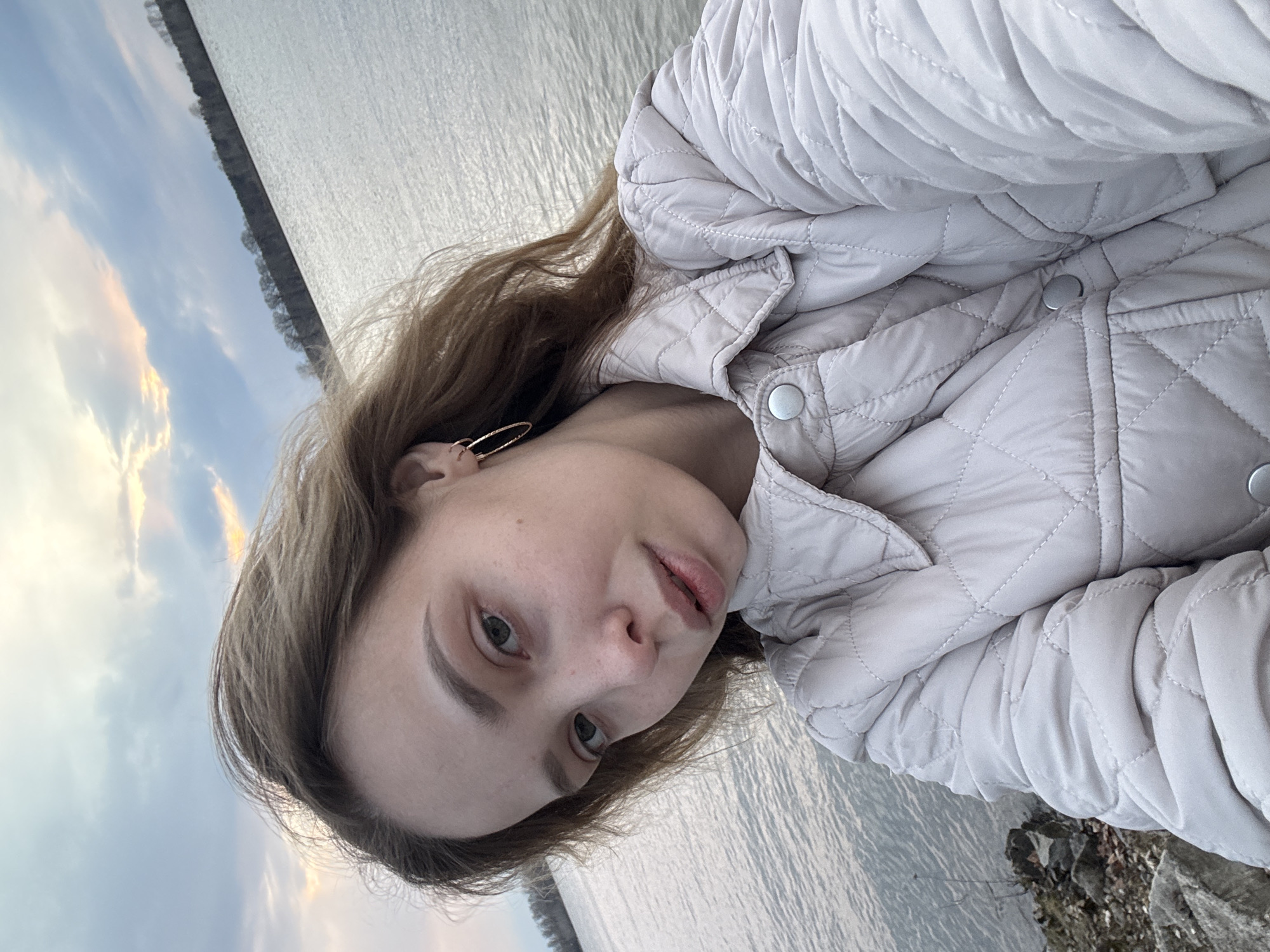 A young woman is depicted in a selfie, with the image rotated 90 degrees clockwise. She is positioned outdoors against a scenic backdrop of a large body of water and a partly cloudy sky. The woman has light brown hair, light-colored eyes, and a neutral expression as she gazes towards the camera. She is wearing a light-colored, possibly beige or light grey, quilted jacket with a distinctive diamond pattern and snap buttons. A gold-toned hoop earring is visible in her left ear.
Behind her, a broad expanse of water with subtle ripples extends, suggesting a river or lake. A faint line of distant trees or land marks the horizon across the water. The sky above is partly cloudy, featuring a blend of blues, whites, and soft orange or pink tones, indicating either sunrise or sunset. The light is soft and diffused. In what would be the lower right corner if the image were upright, some rocky terrain is visible at the water's edge. This natural setting, featuring water and sky, is consistent with a location such as Noviy, Russia. No text is visible in the image.